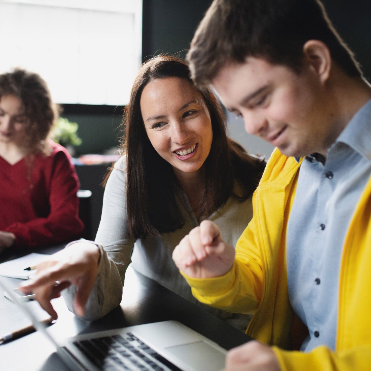 Woman and man with Down syndrome look at laptop, smiling. A third person is in the background.
