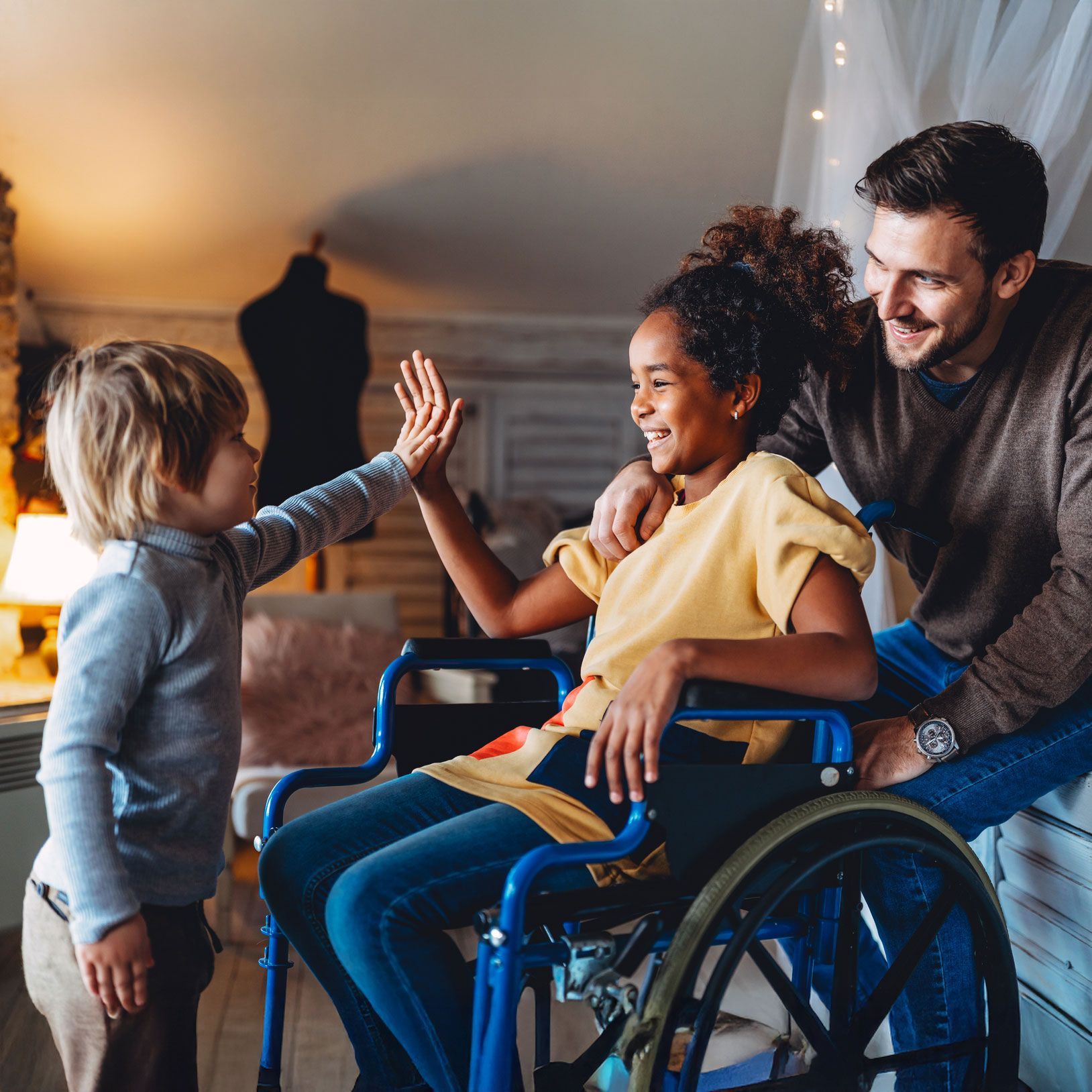 A smiling girl in a wheelchair high-fives a boy while a man smiles, indoors, warm lighting.