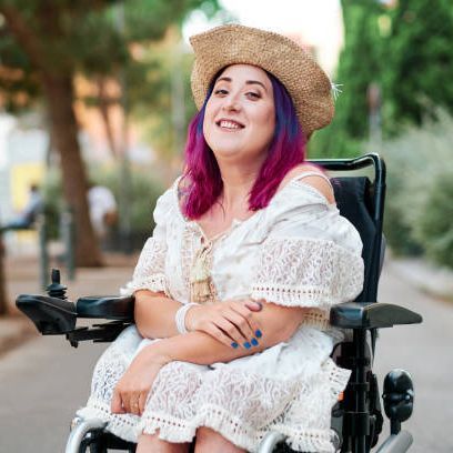 Woman in a straw hat and white dress smiles while sitting in a wheelchair outdoors.