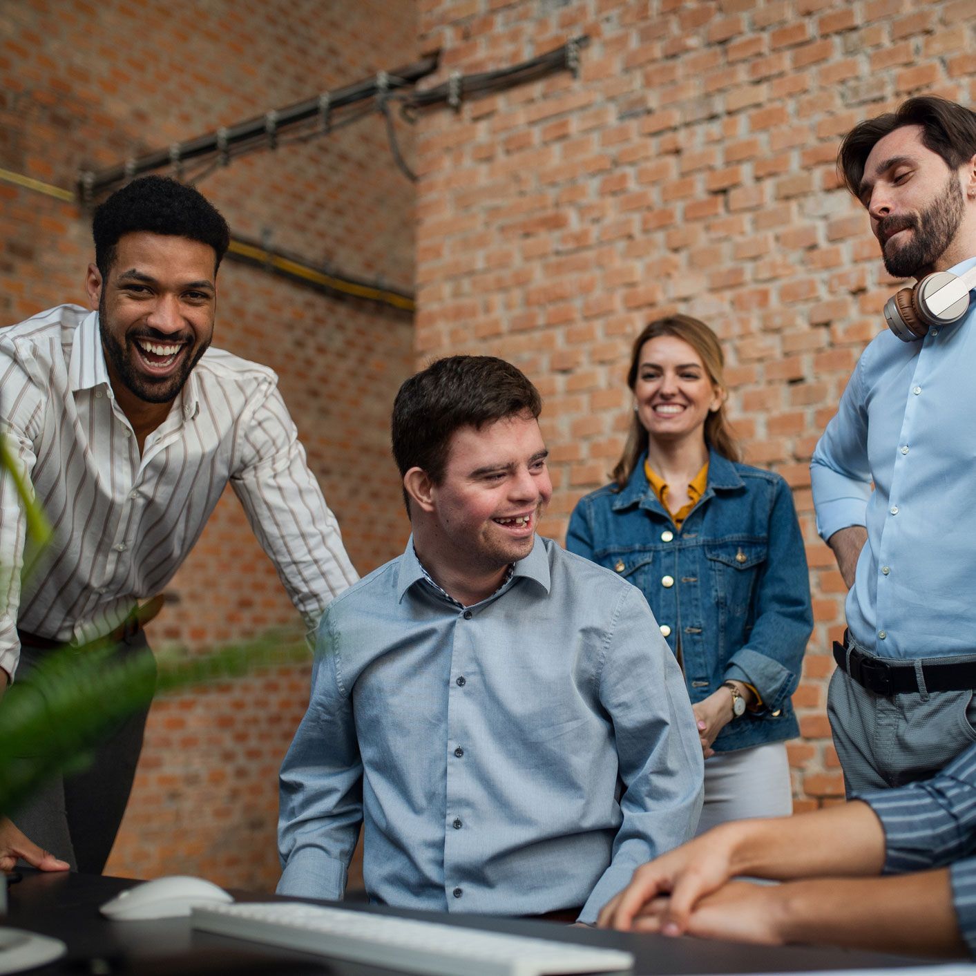 Office team smiles together; brick wall background. Man with Down syndrome centered.