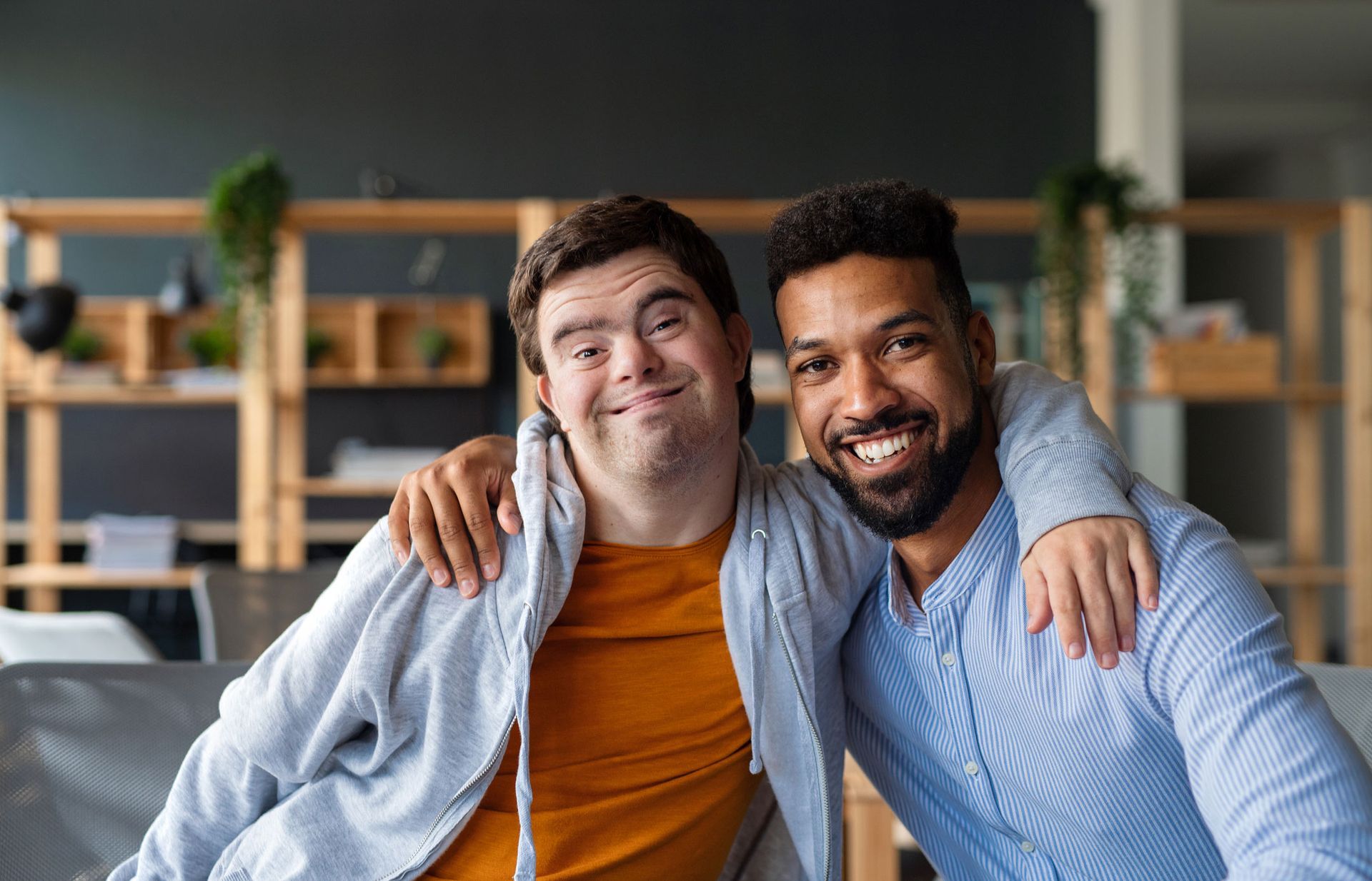 Two smiling men with arms around each other. One has Down syndrome; they are indoors in a casual setting.