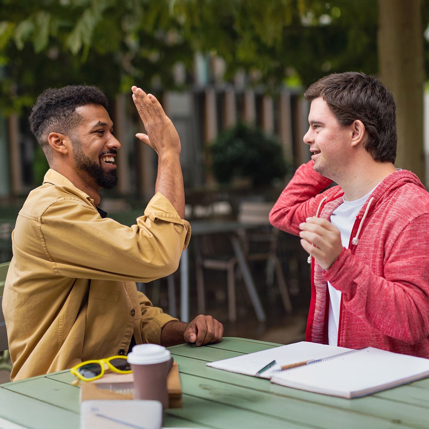 Man high-fiving another man with Down syndrome at a table outdoors. Both smile.