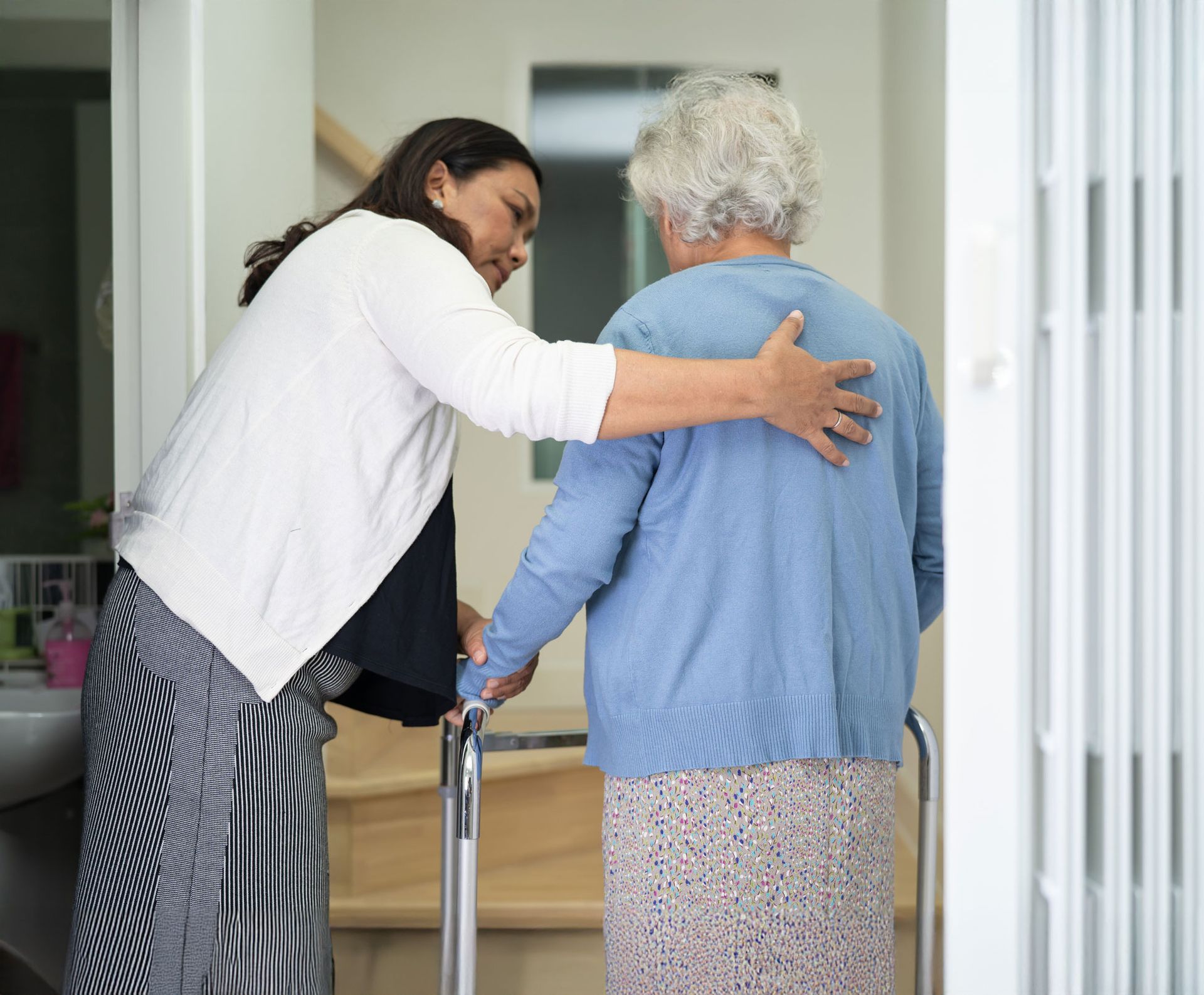 Woman helping elderly woman with a walker; home interior, stairs in background.