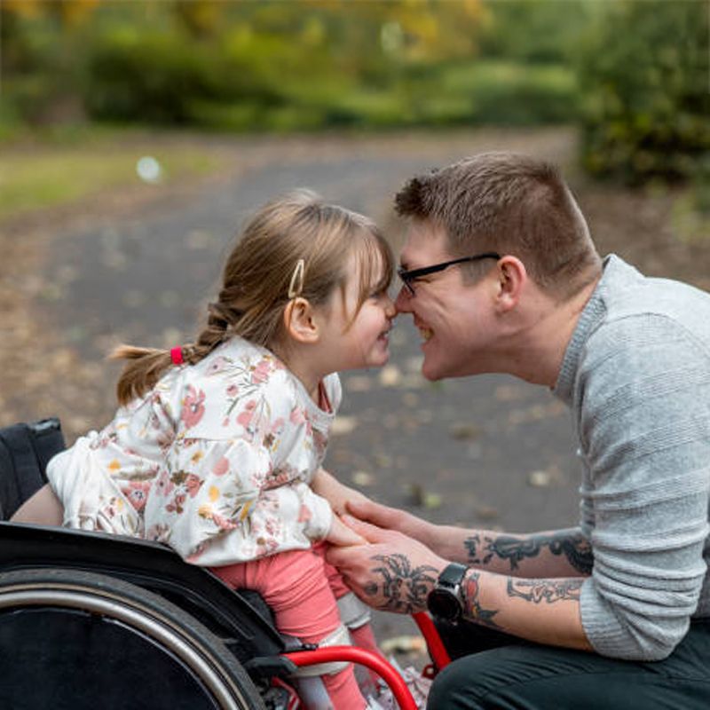Man and young girl in wheelchair touch noses on path; outdoors.