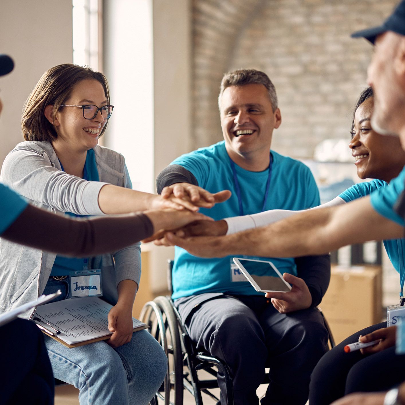 Group of volunteers, some with disabilities, putting their hands together in a circle, smiling indoors.