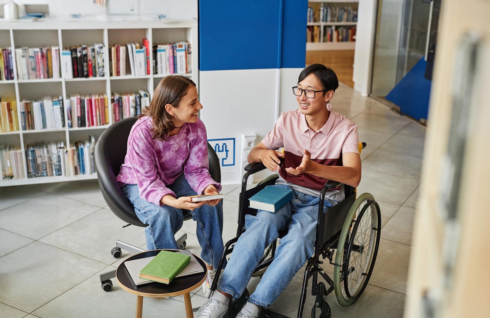 Woman and man in wheelchair chat in a library. They are smiling, surrounded by books.