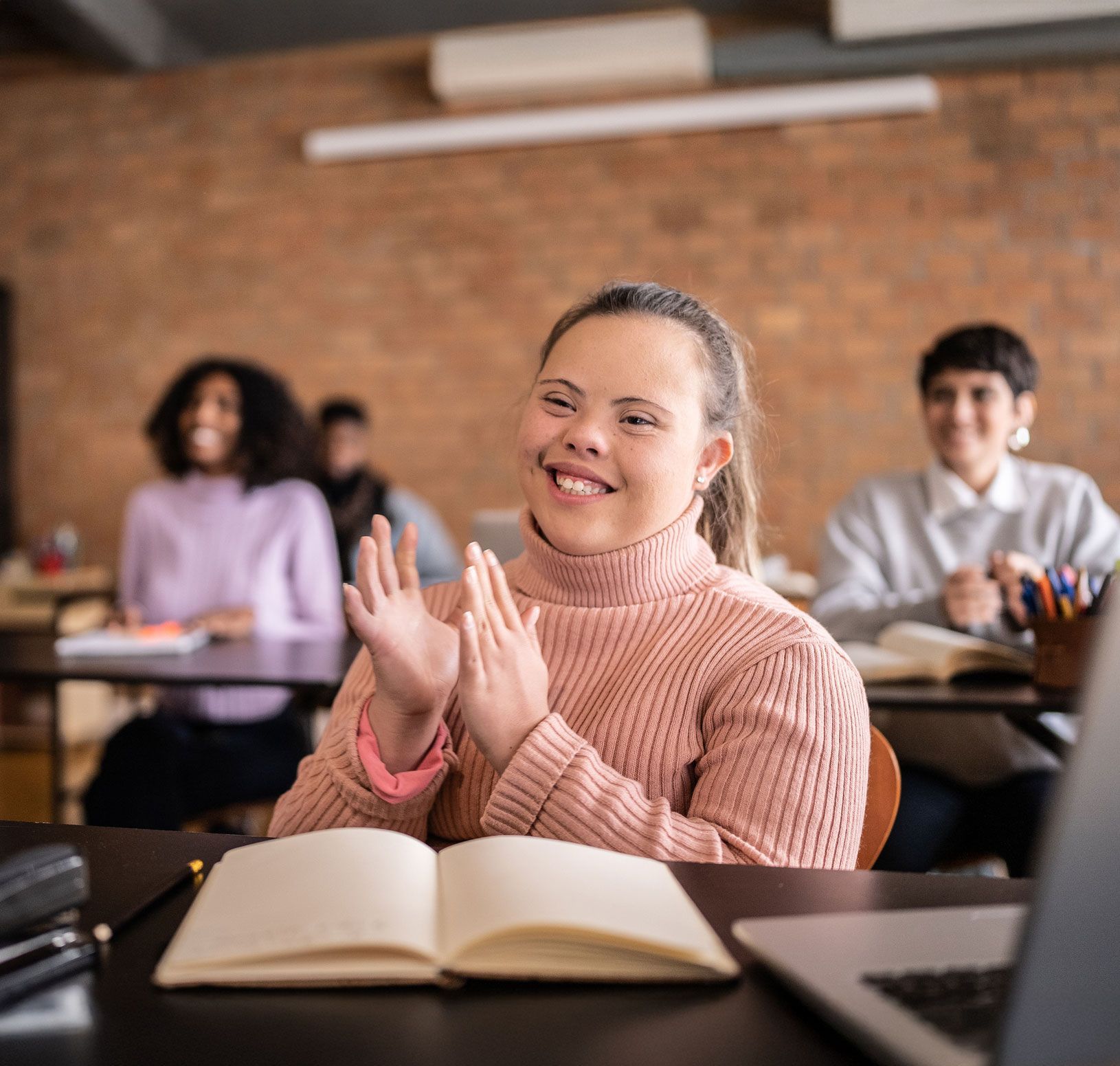 Woman with Down syndrome clapping enthusiastically in a classroom, smiling, with classmates in the background.