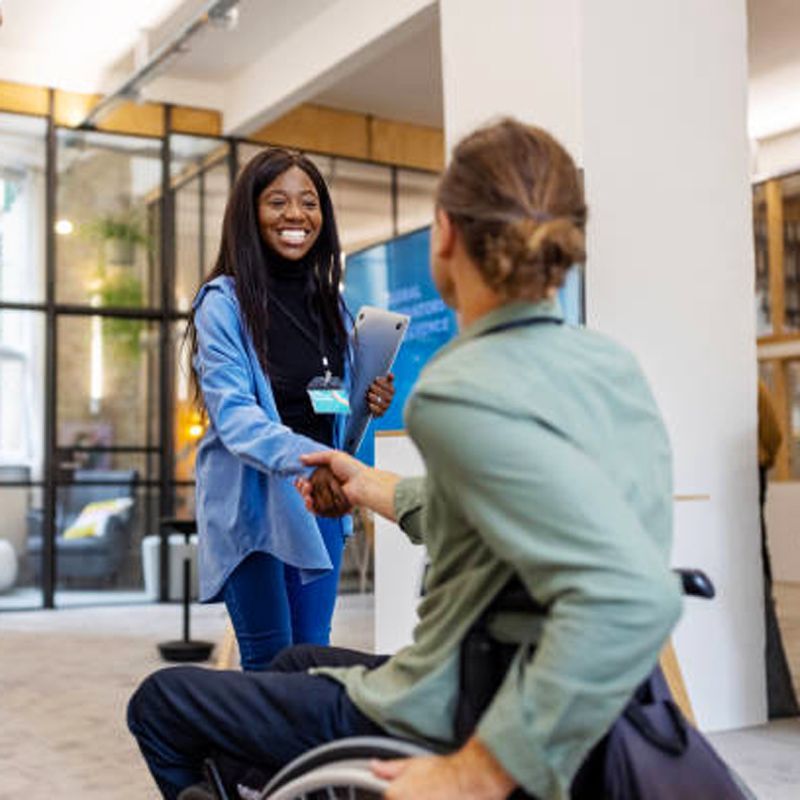 Woman in blue shakes hands with a man in a wheelchair indoors, smiling.