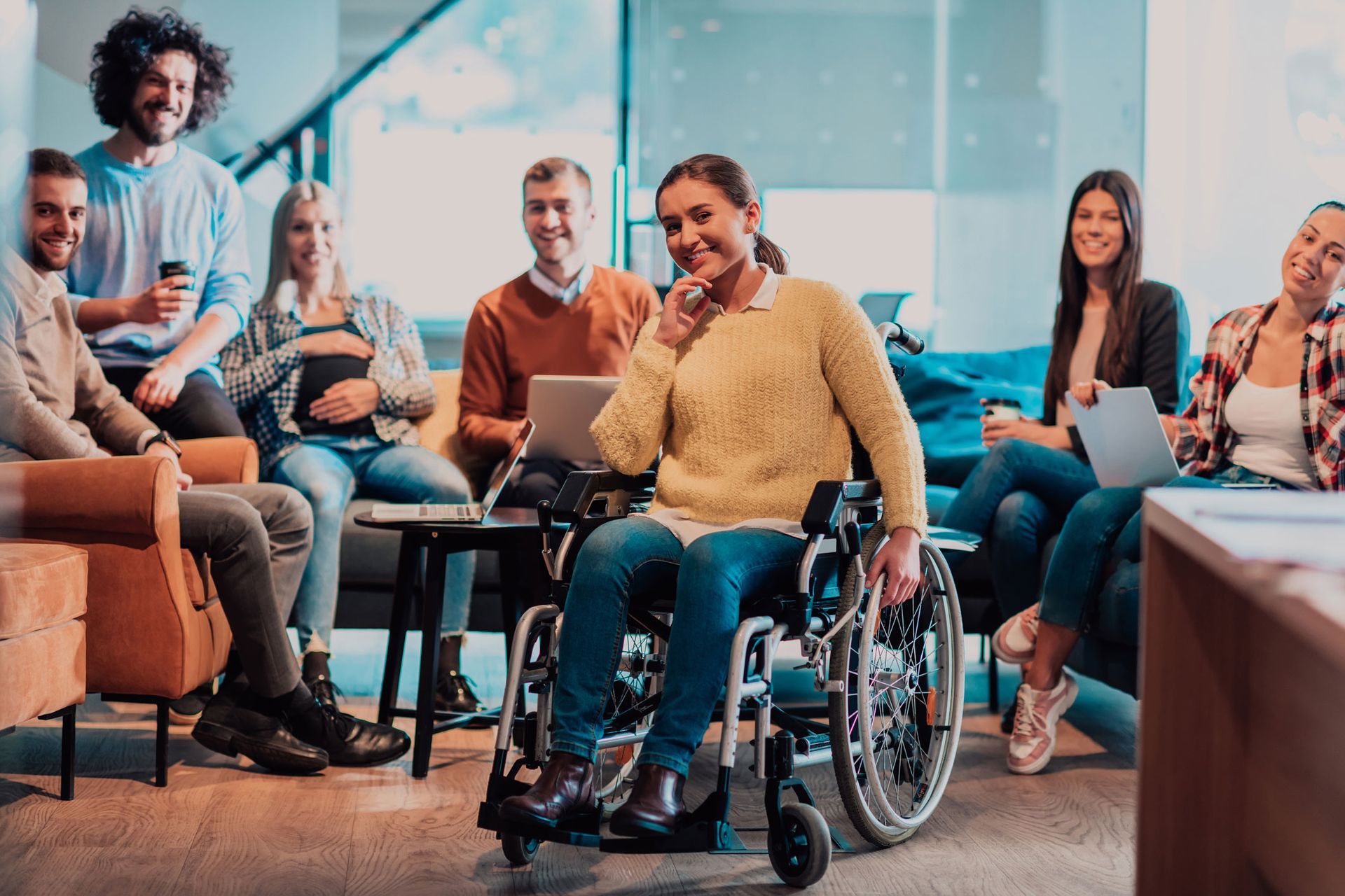 Group of people in a modern office, woman in wheelchair smiles, others work on laptops, relaxed and collaborative.