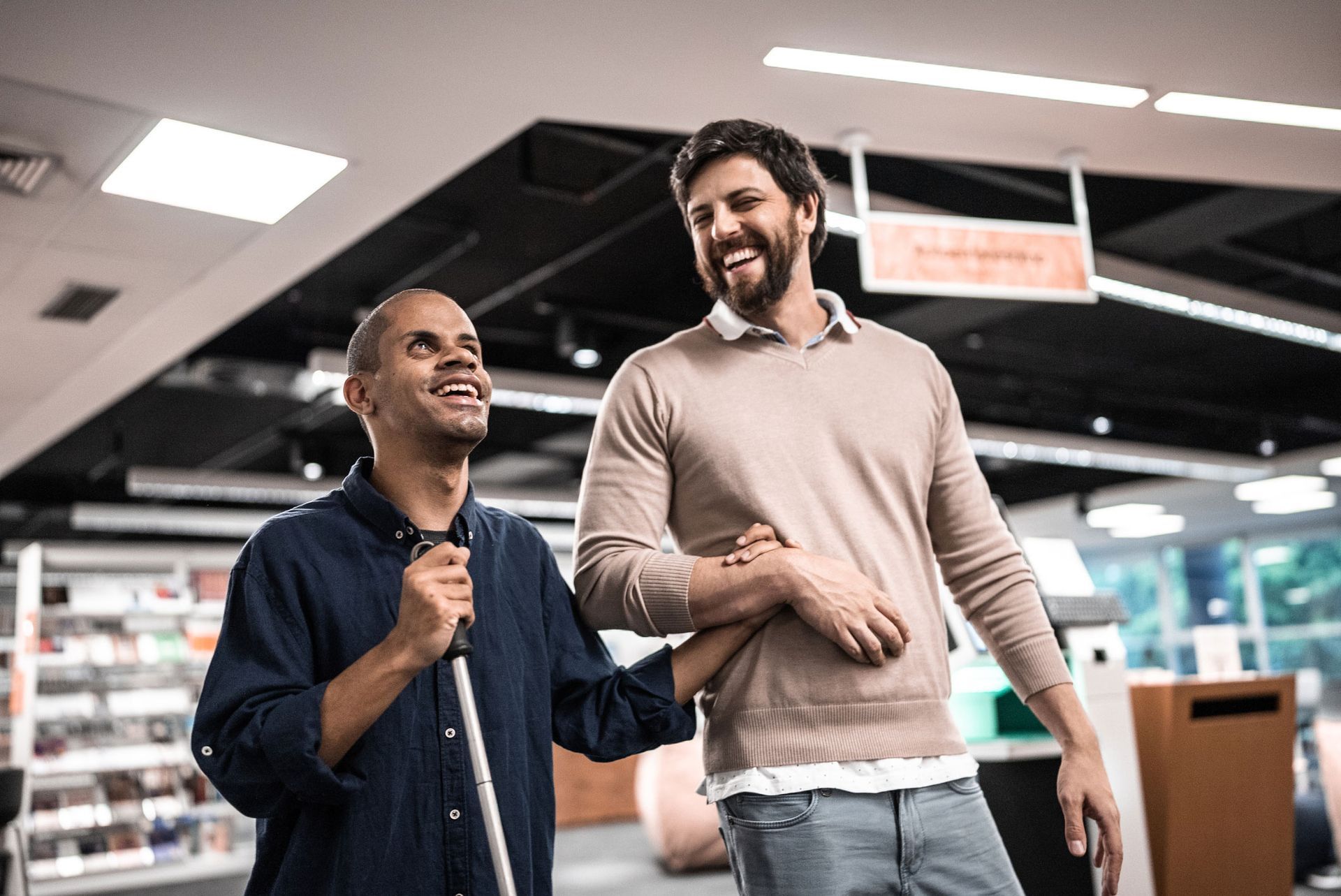 Man with cane and another man arm in arm, smiling while walking in a store.