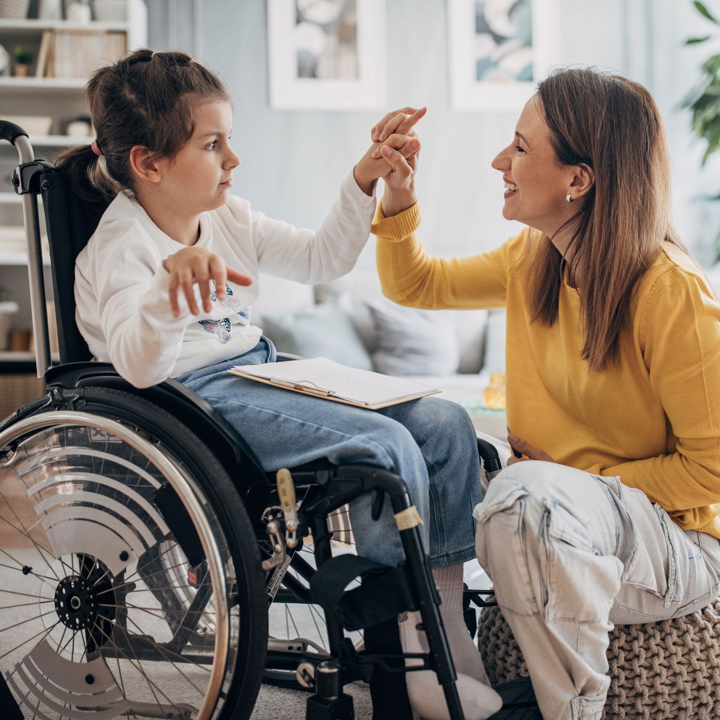 A child in a wheelchair high-fives a smiling woman indoors.