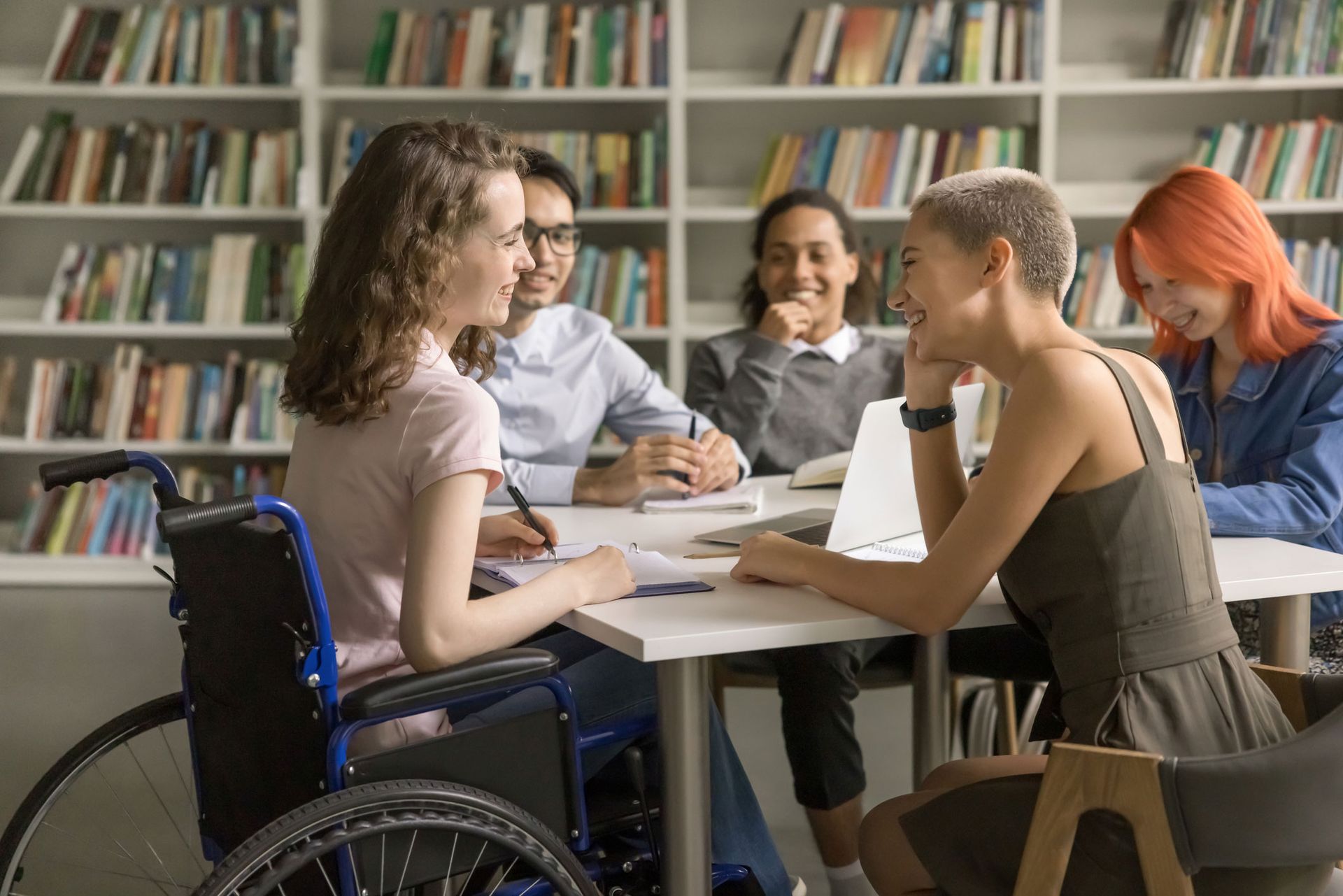 Group of diverse students collaborating at a table in a library; one student uses a wheelchair.