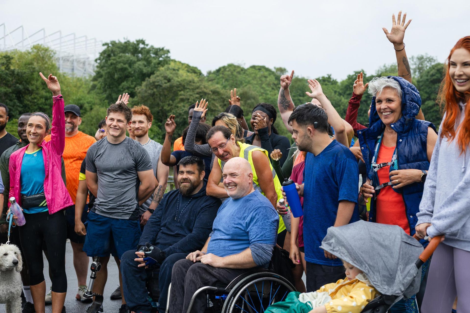 Group of diverse people, some in wheelchairs, cheering with arms raised outdoors. Park setting, cloudy day.