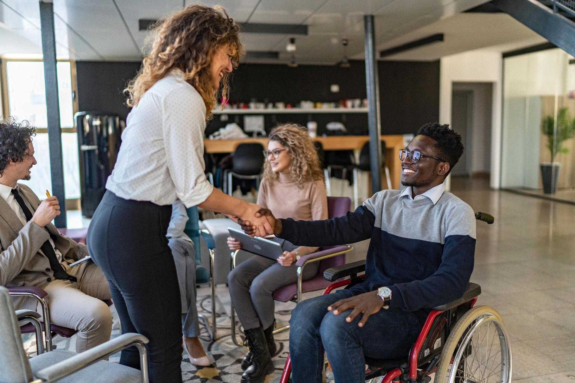 Woman in white shirt shaking hands with Black man in wheelchair; colleagues watch in a modern office.