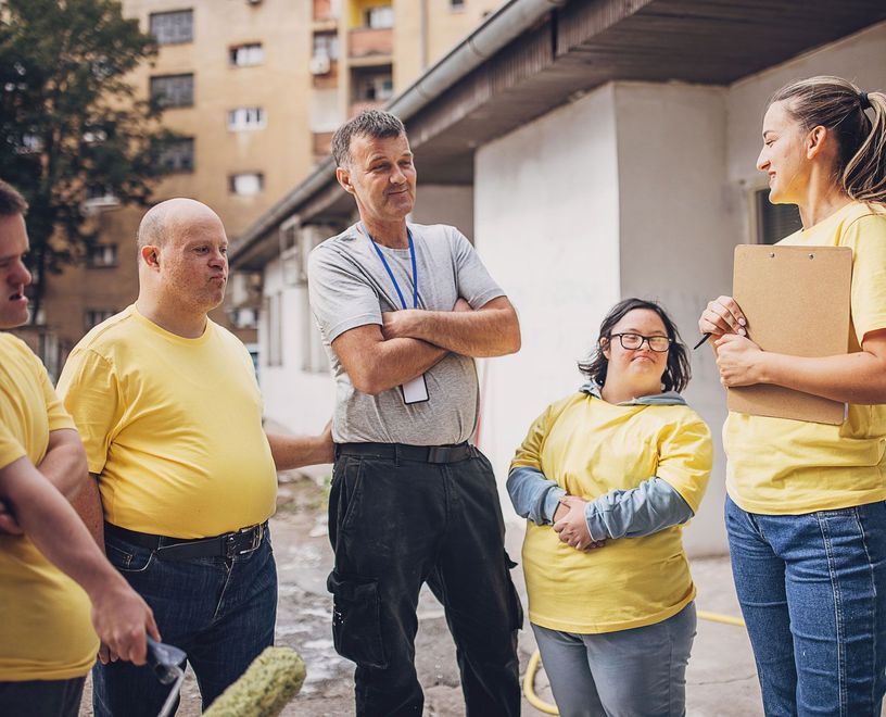 Volunteers in yellow shirts and a man with a lanyard work on a building. Woman with notepad. Outdoors.