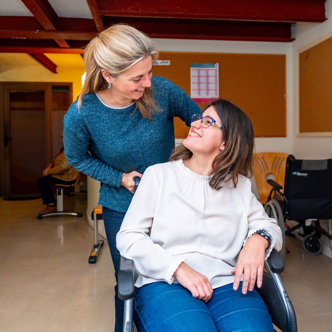 Woman assisting another woman in a wheelchair, both smiling, indoors.