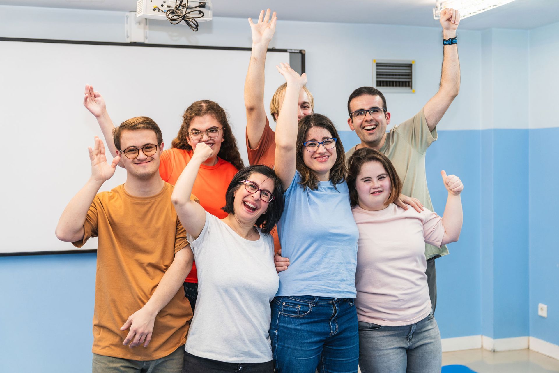Group of diverse adults happily raising arms in a room with a whiteboard and blue walls.