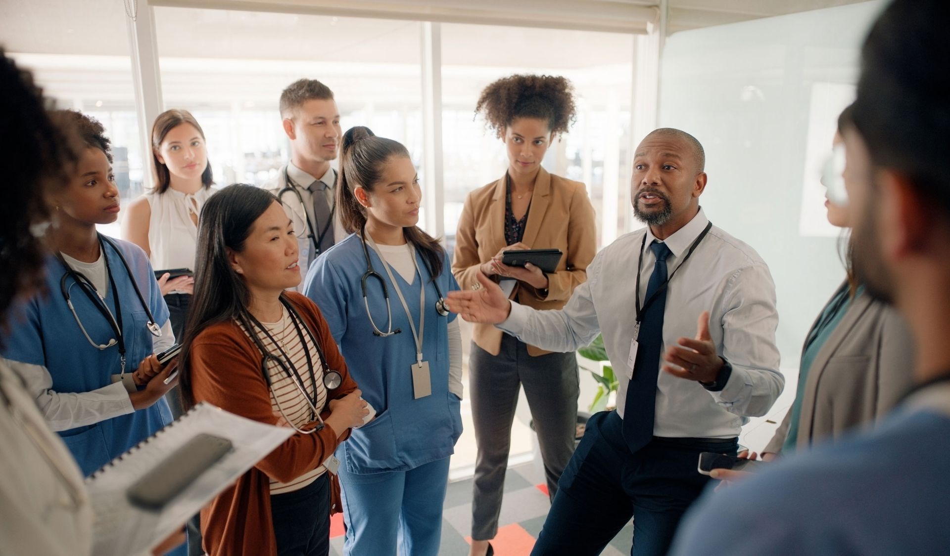 A diverse group of medical professionals in a bright office, listening to a Black man speaking with hands gesturing.