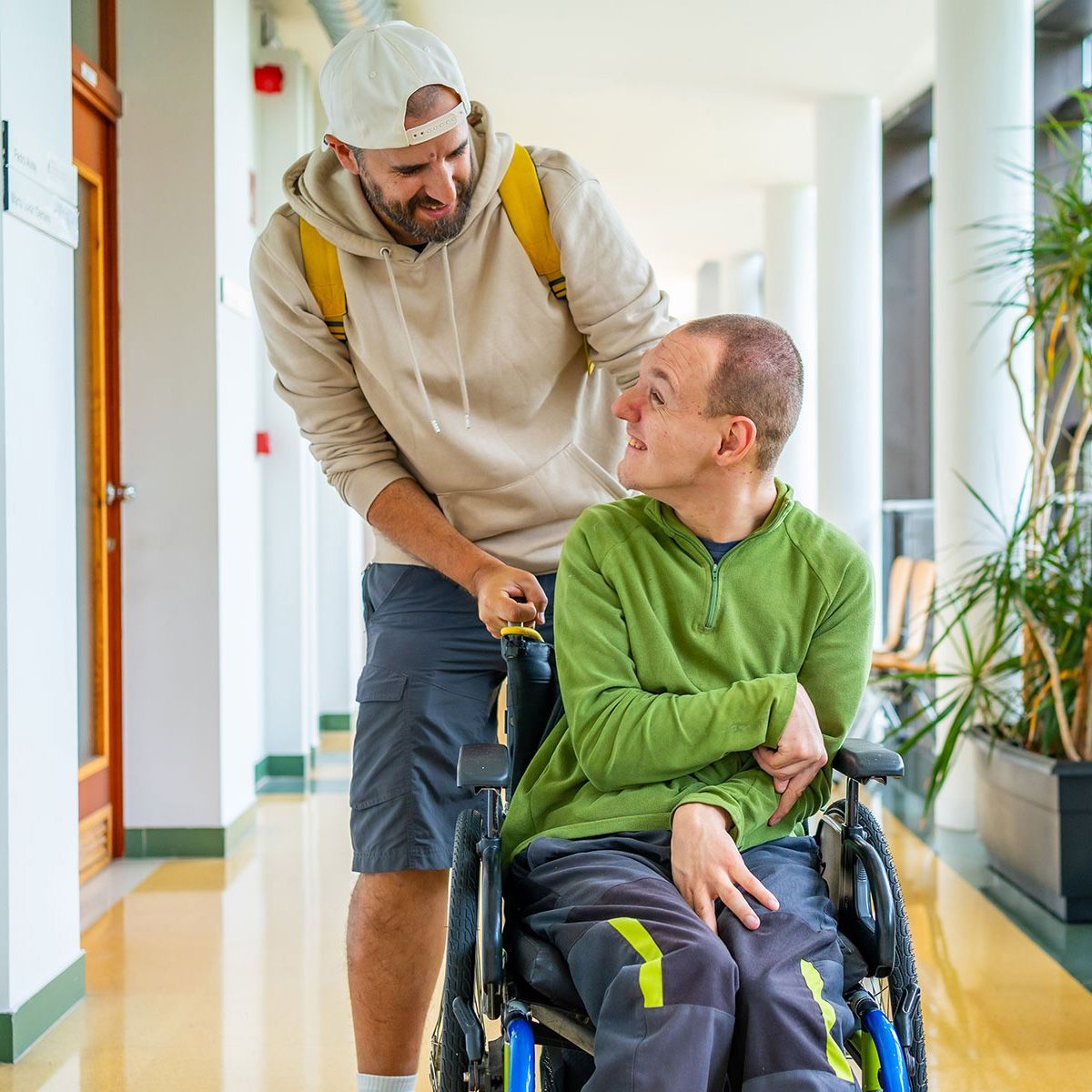 Man in wheelchair is pushed by another man in a hallway; both men smile.