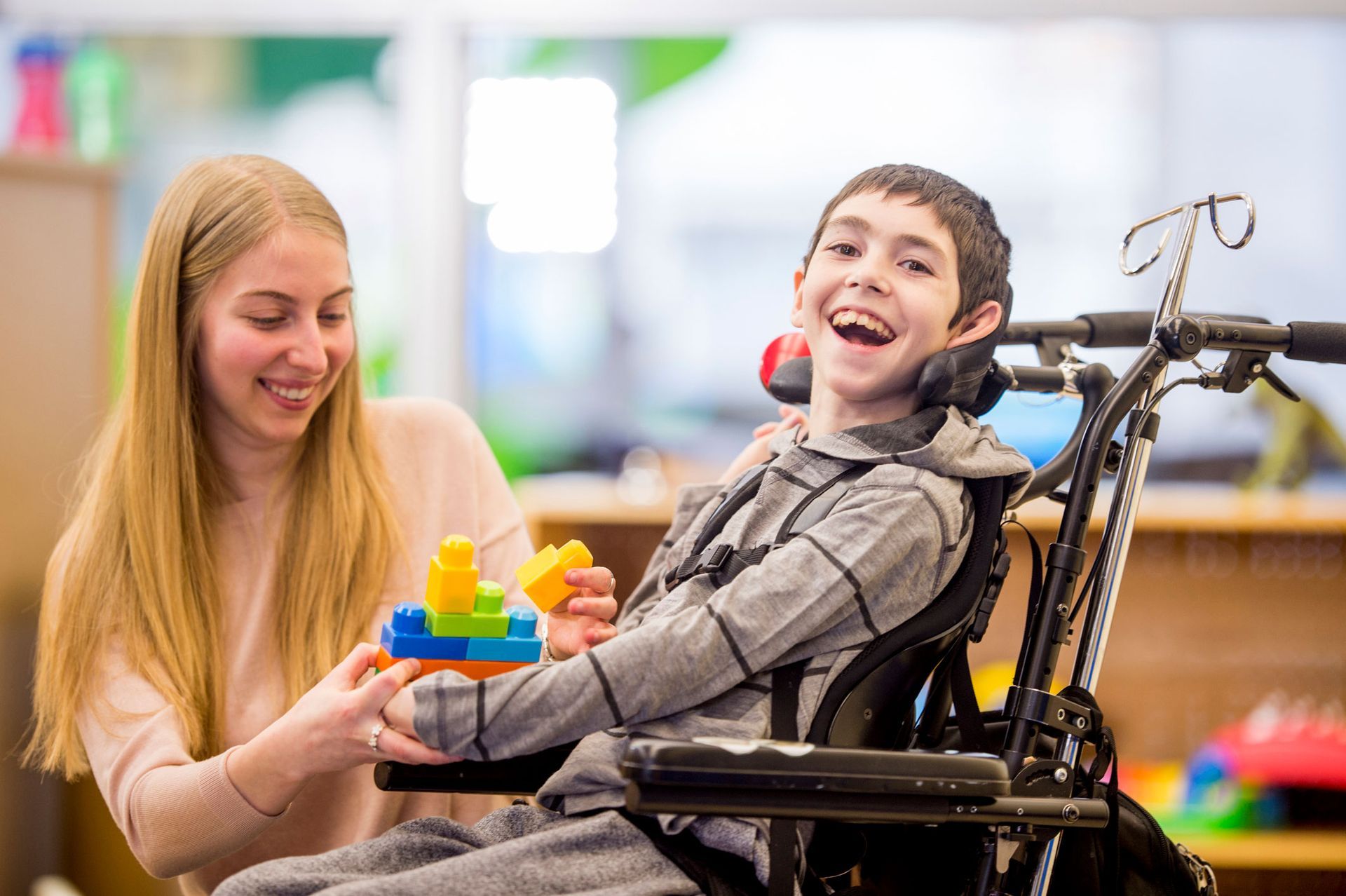 Woman helps a smiling boy in a wheelchair build a toy tower; indoors.