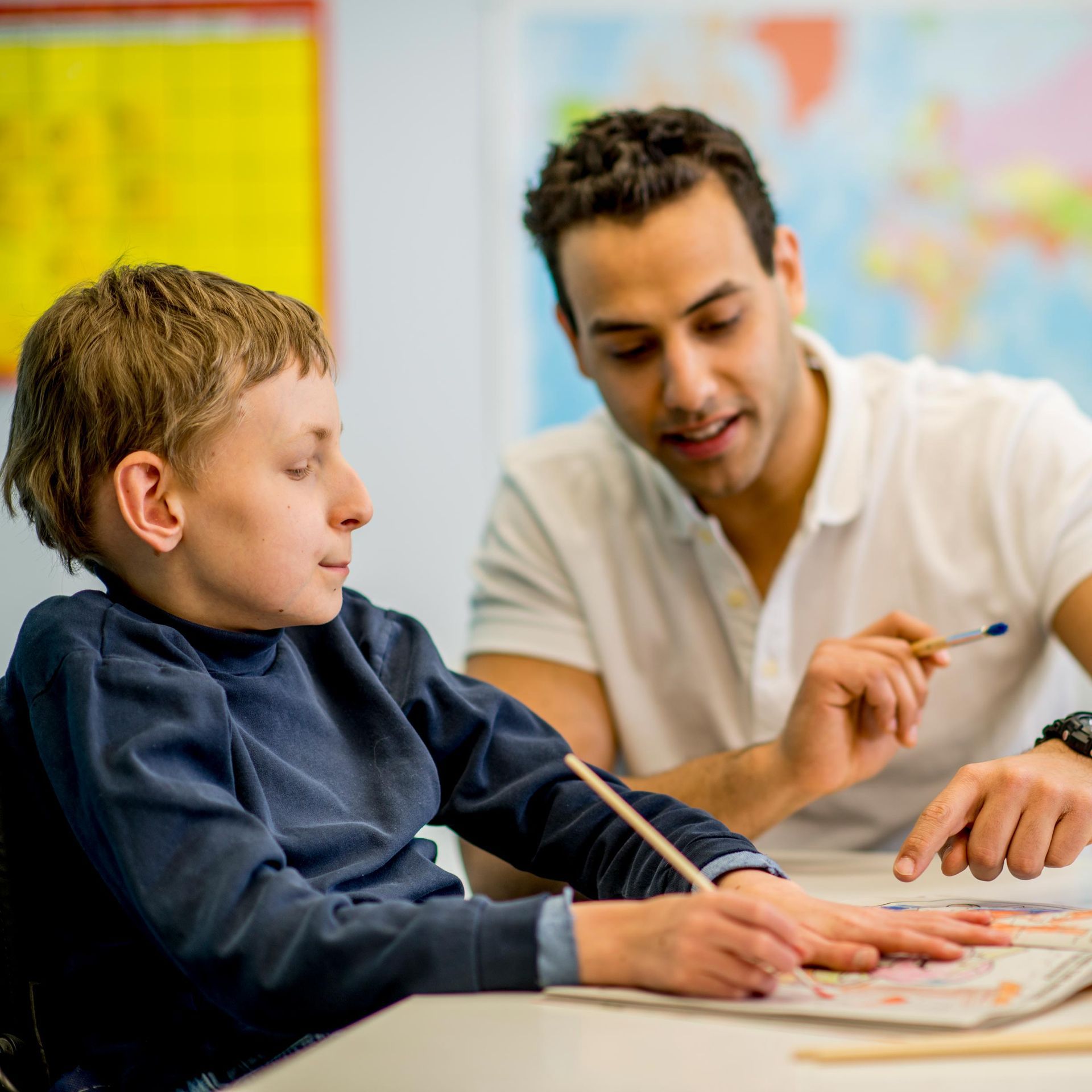 A young man assists a student with drawing at a desk.  Both are smiling.