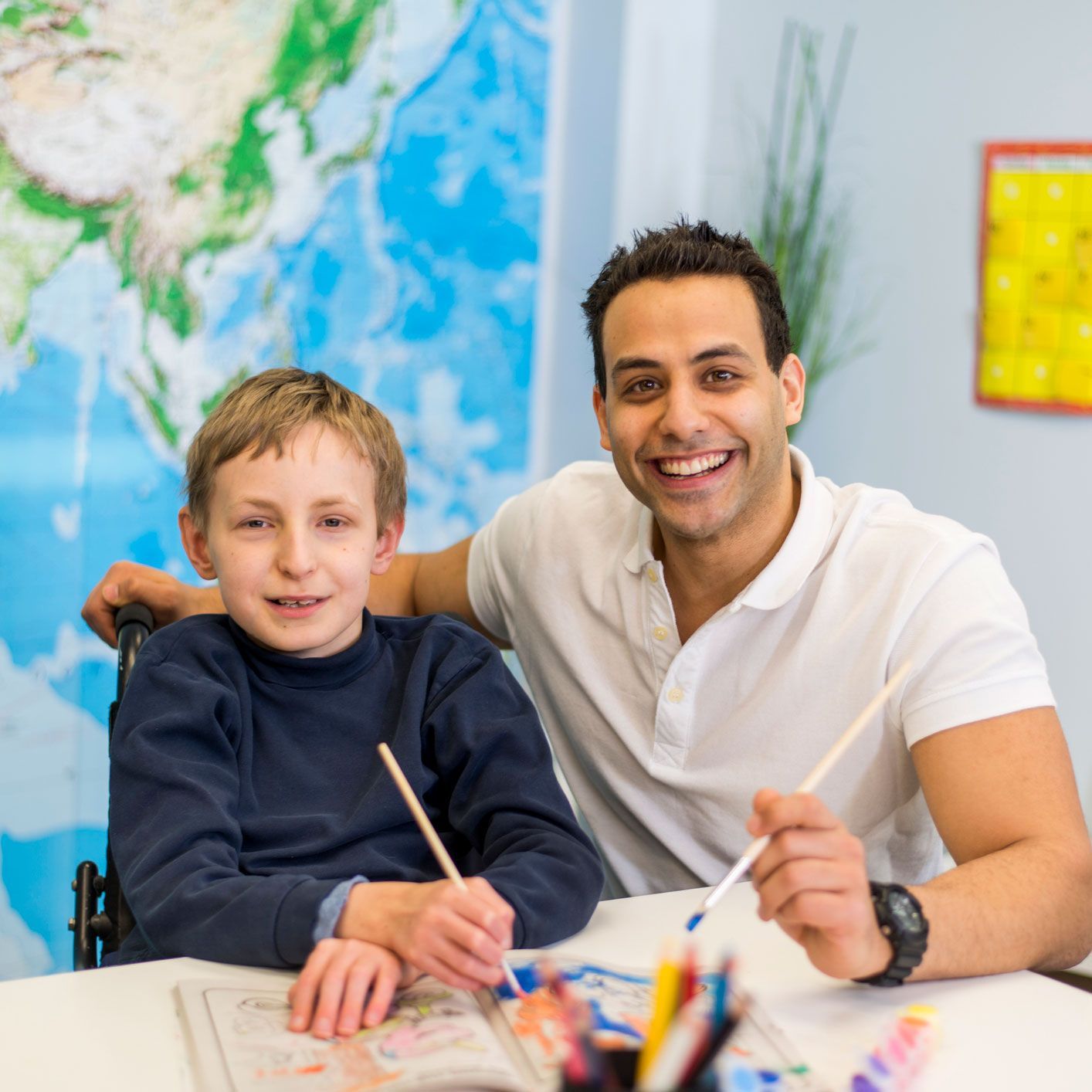 Smiling man with boy in wheelchair painting at a table; world map in background.