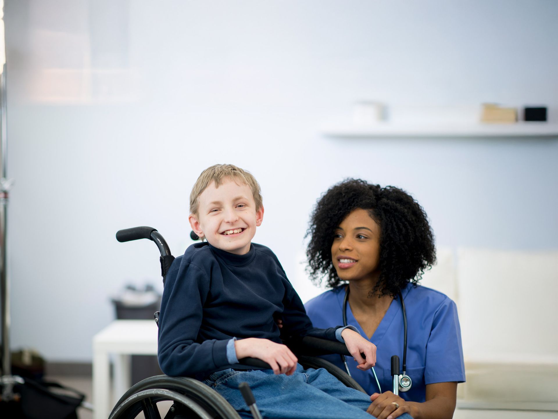 Boy in wheelchair smiling at the camera; nurse in blue scrubs smiles next to him.