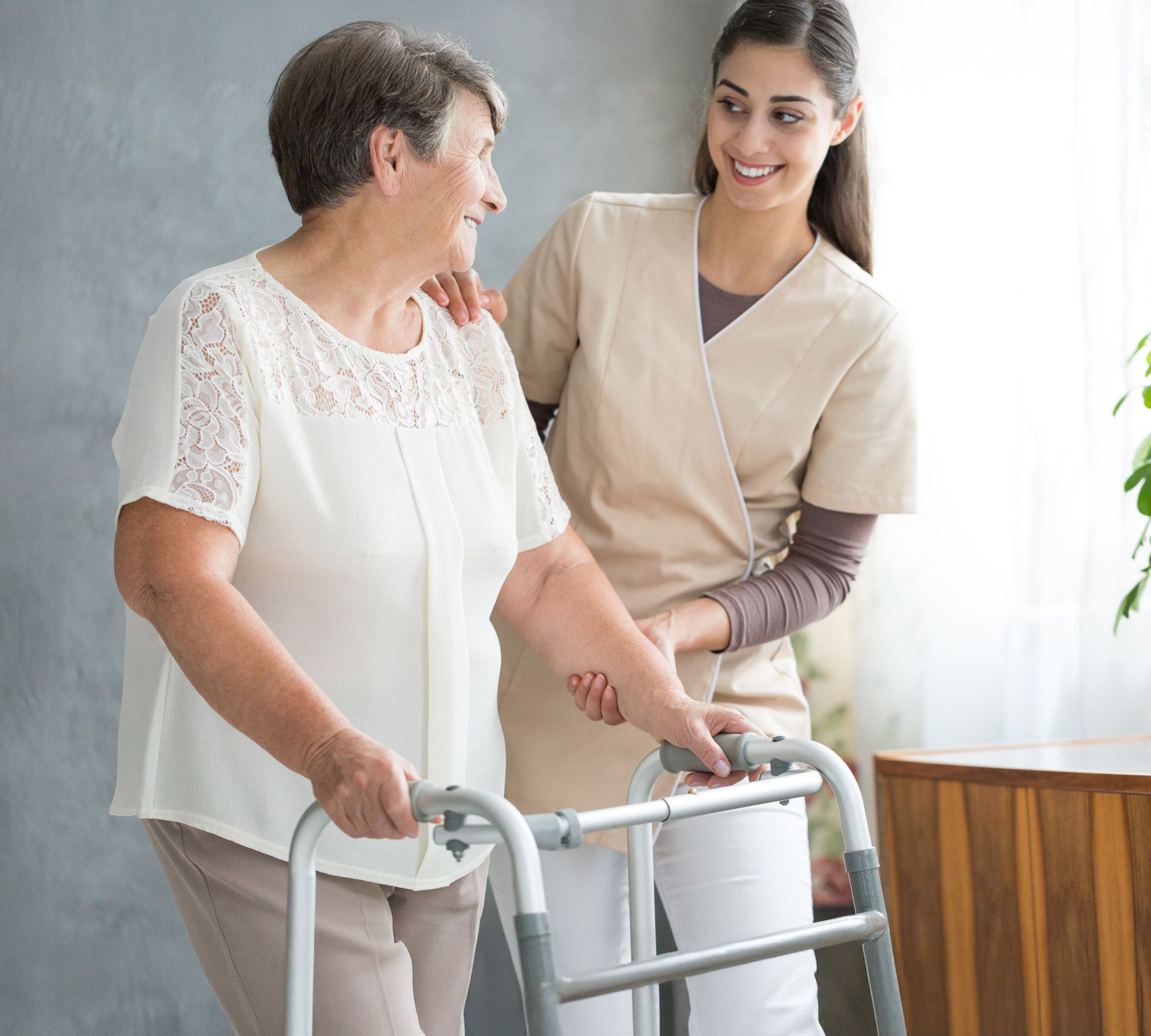 An older woman uses a walker with assistance from a smiling caregiver, indoors.