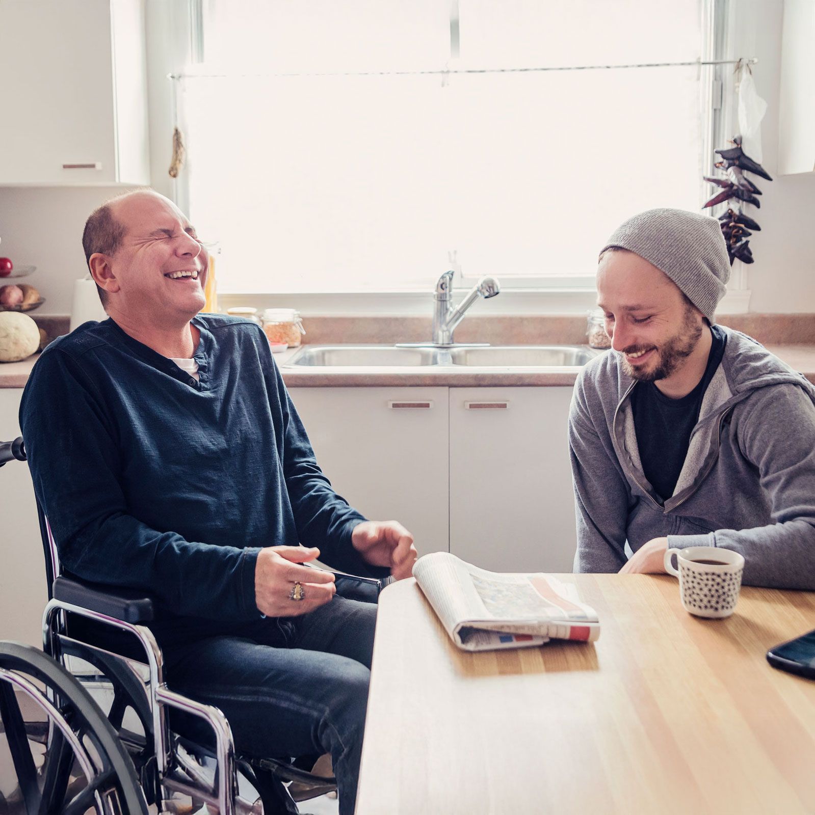 Man in wheelchair laughing with another man at a kitchen table; newspaper and coffee cup present.