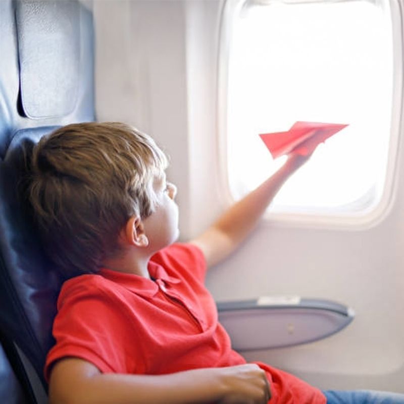 Boy on an airplane holding a red paper airplane near the window.