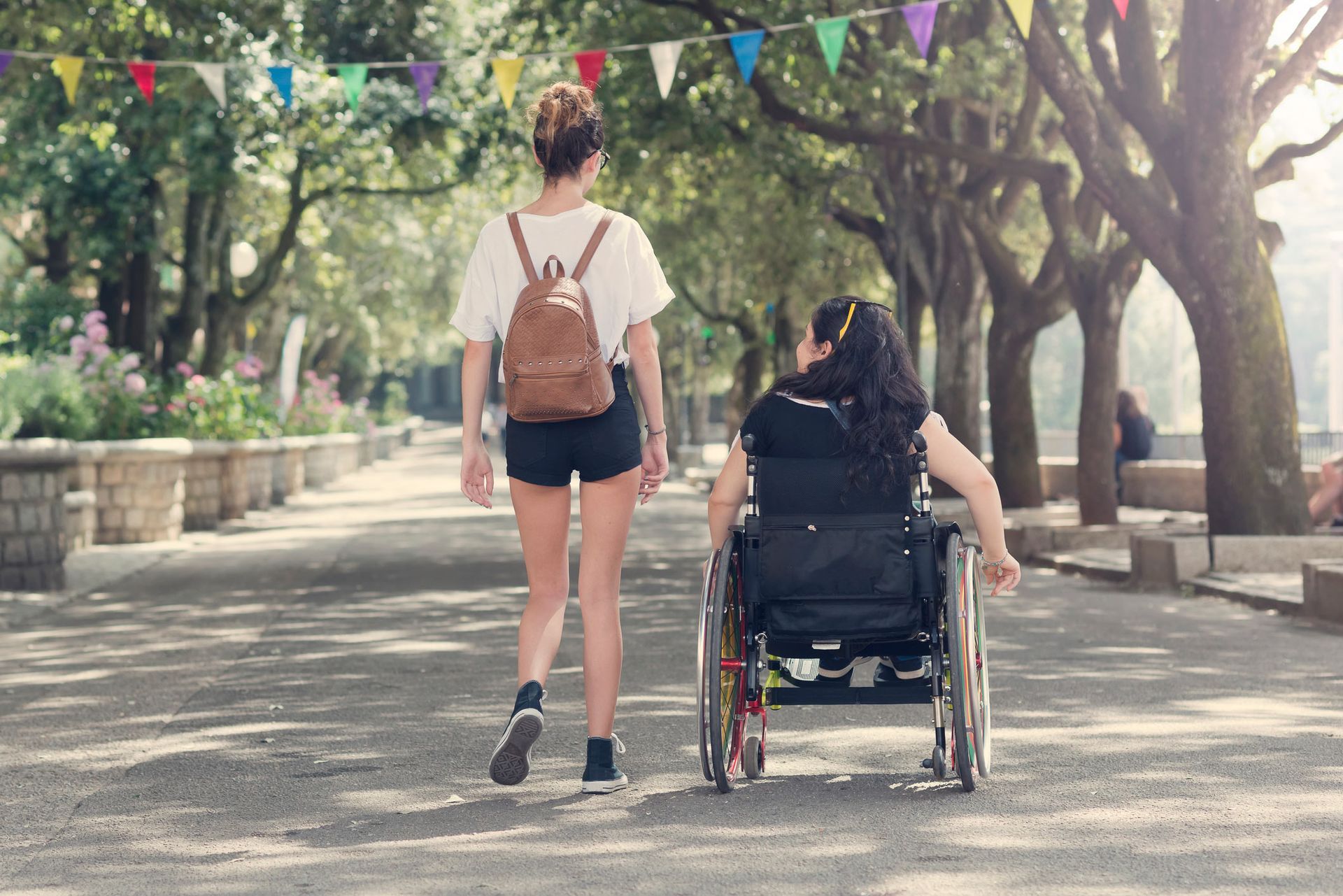 A woman in a wheelchair and a woman walking together on a path in a park, sunny day.