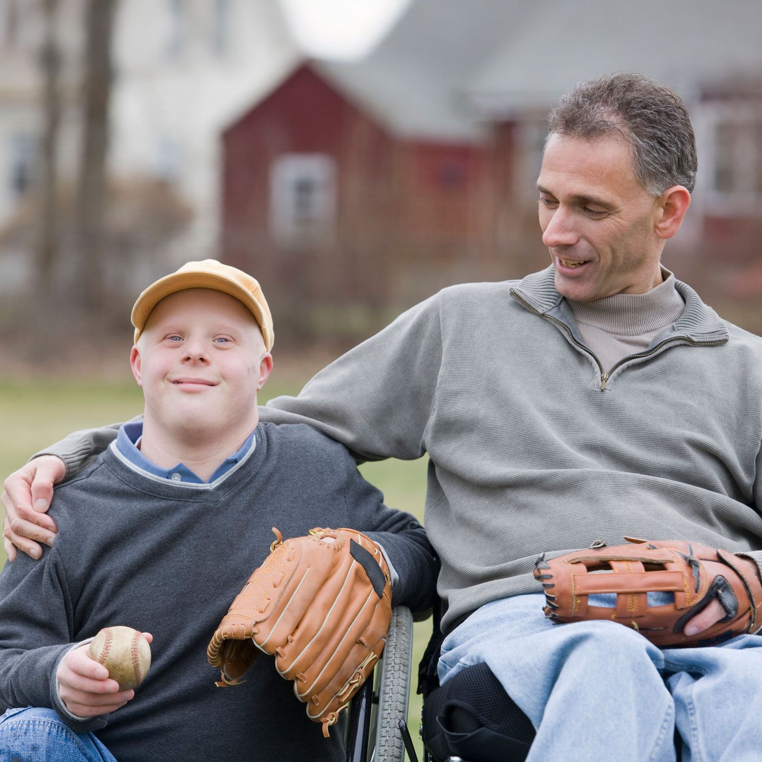 A smiling boy in a wheelchair holds a baseball and mitt next to a man with his arm around him, in front of a house.
