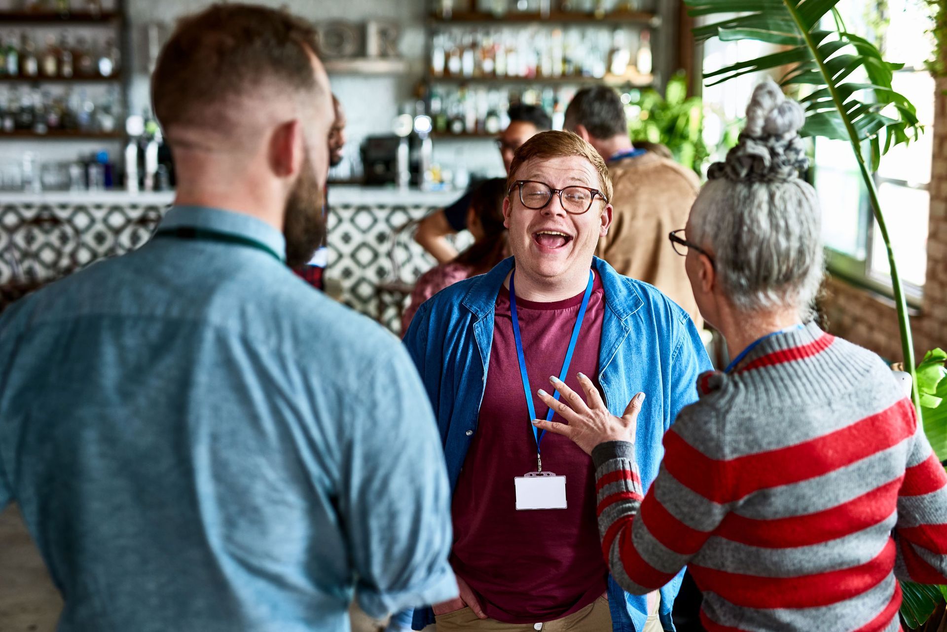 People laughing and talking at an event with name tags, bar in background, and potted plant.