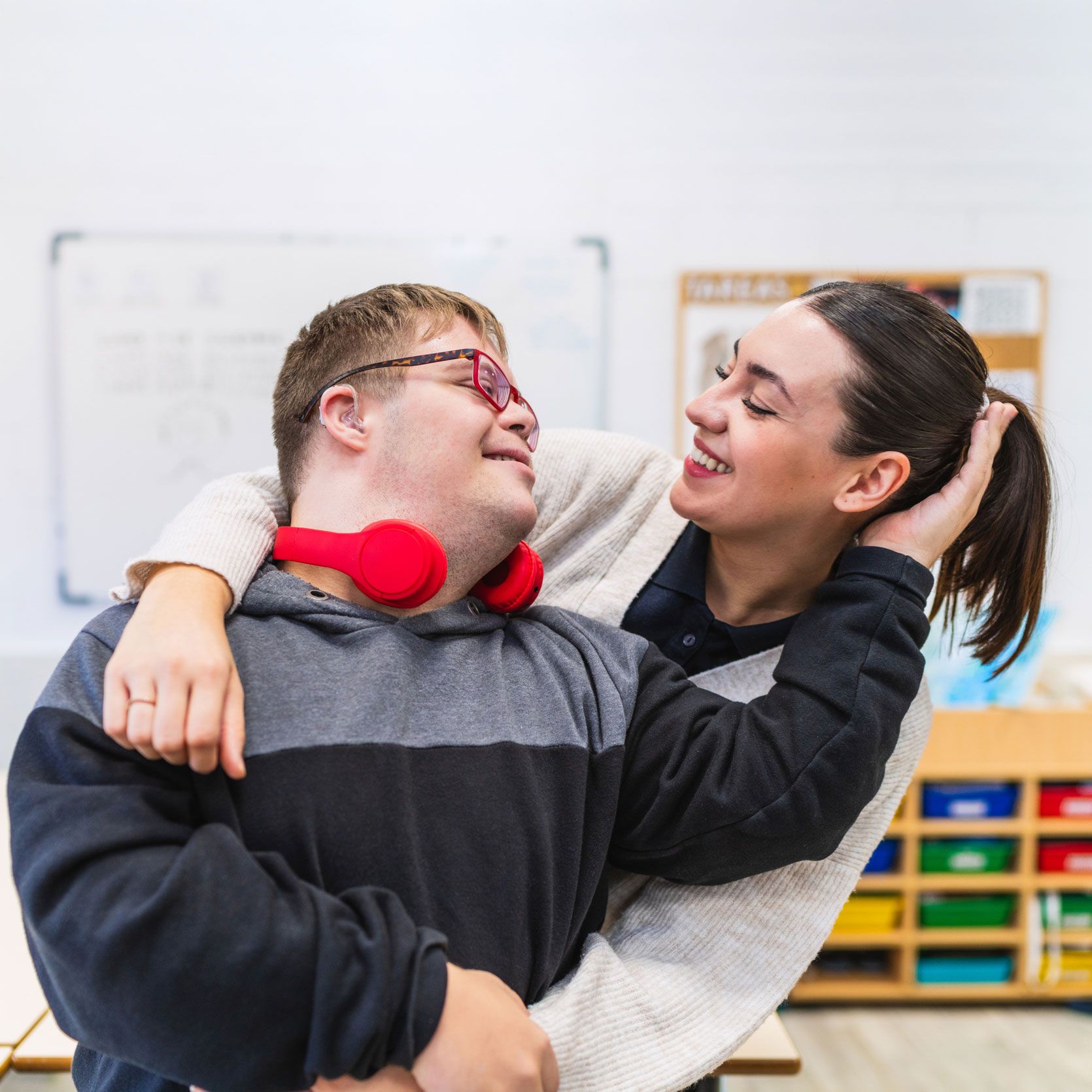 Woman hugs a man with Down syndrome indoors; both smiling. Red headphones around his neck.