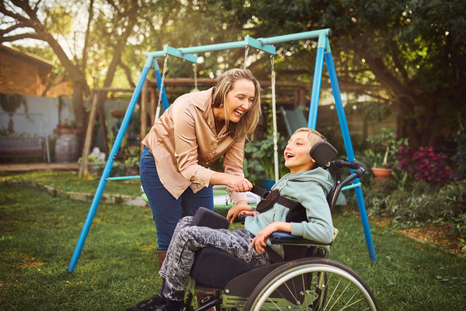 Woman helps child in a wheelchair at a backyard swing set. They are smiling, surrounded by greenery.