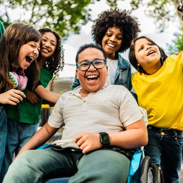 Group of diverse children laughing together, one in a wheelchair. Outdoor setting, bright colors.