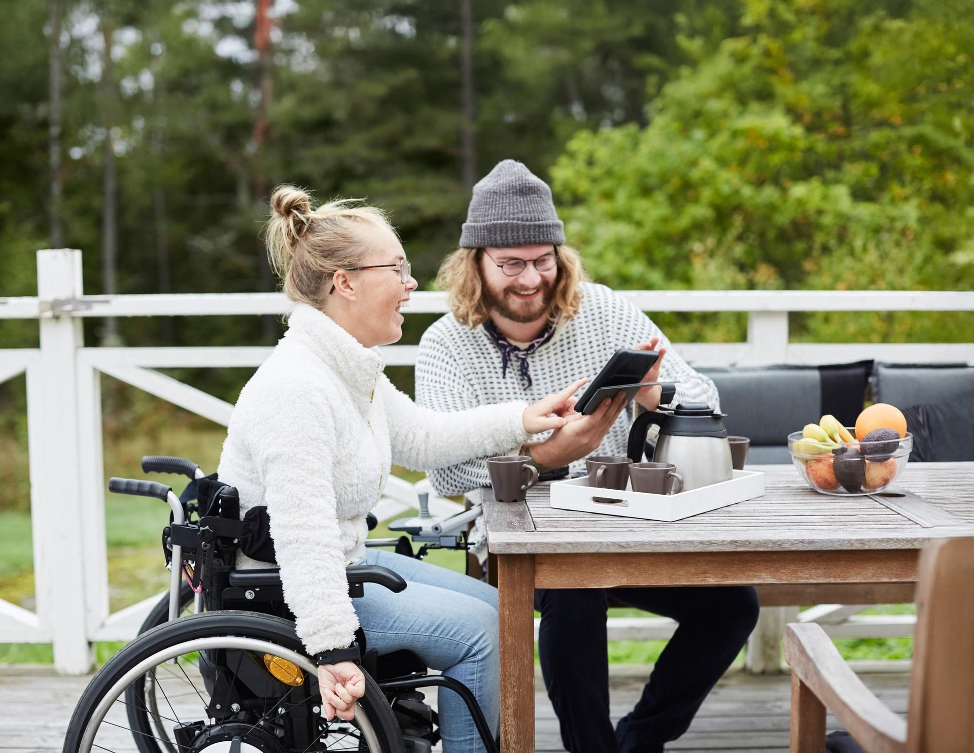 Woman in wheelchair and man look at a tablet on a deck; coffee, fruit, and greenery nearby.