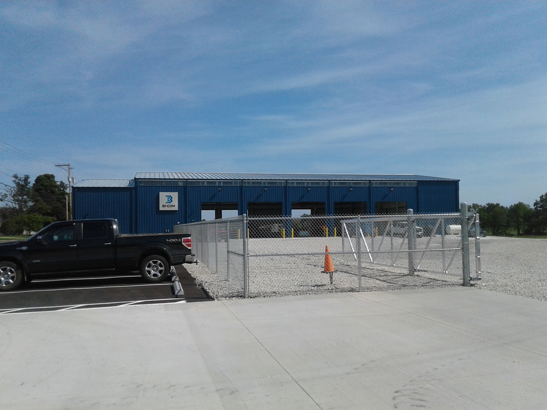 A blue building behind a chain-link fence, with a black truck parked to the left. Sunny, outdoor setting.