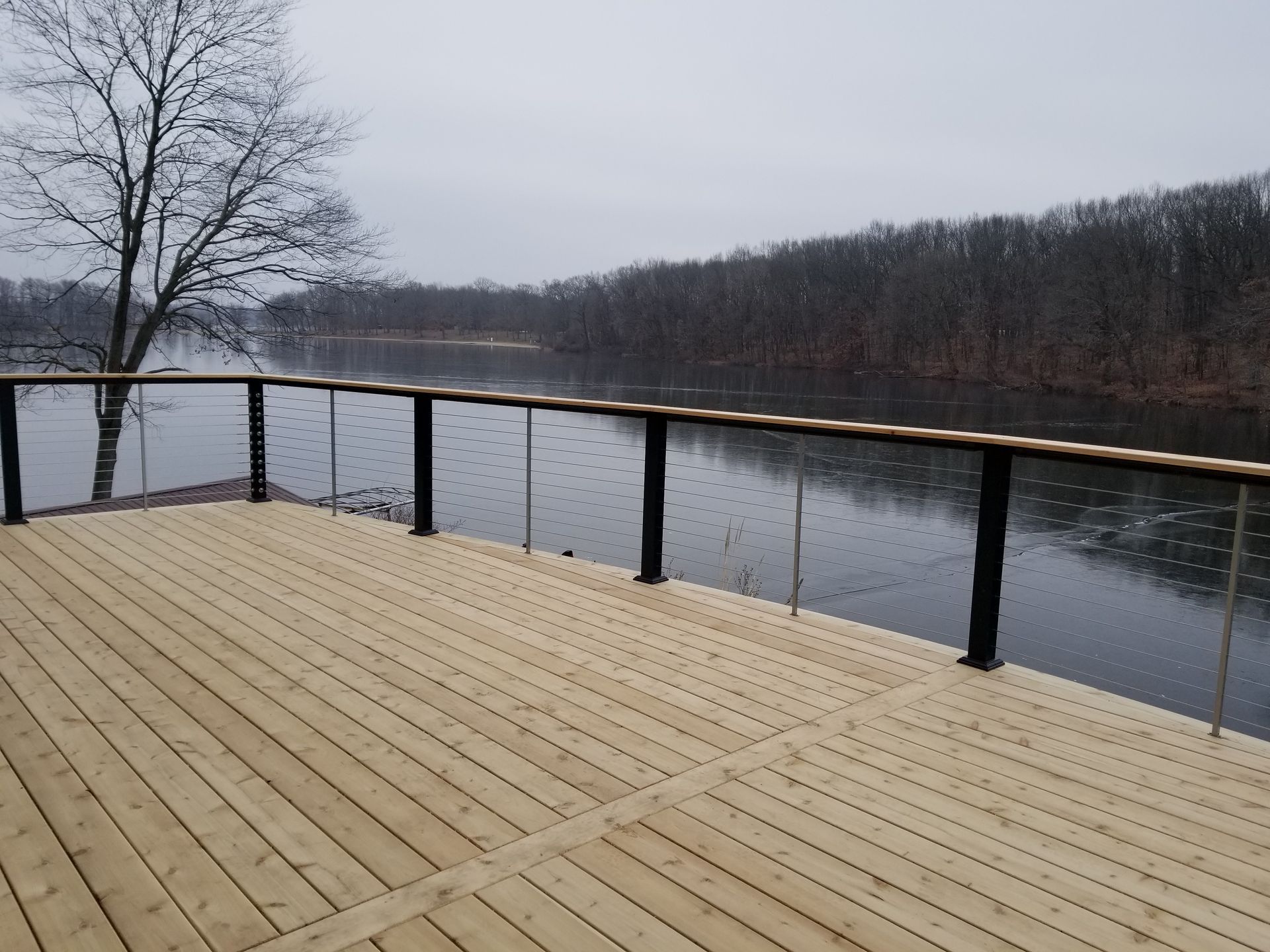 Wooden deck with black railing and cable overlooking a calm river surrounded by bare trees under a cloudy sky.