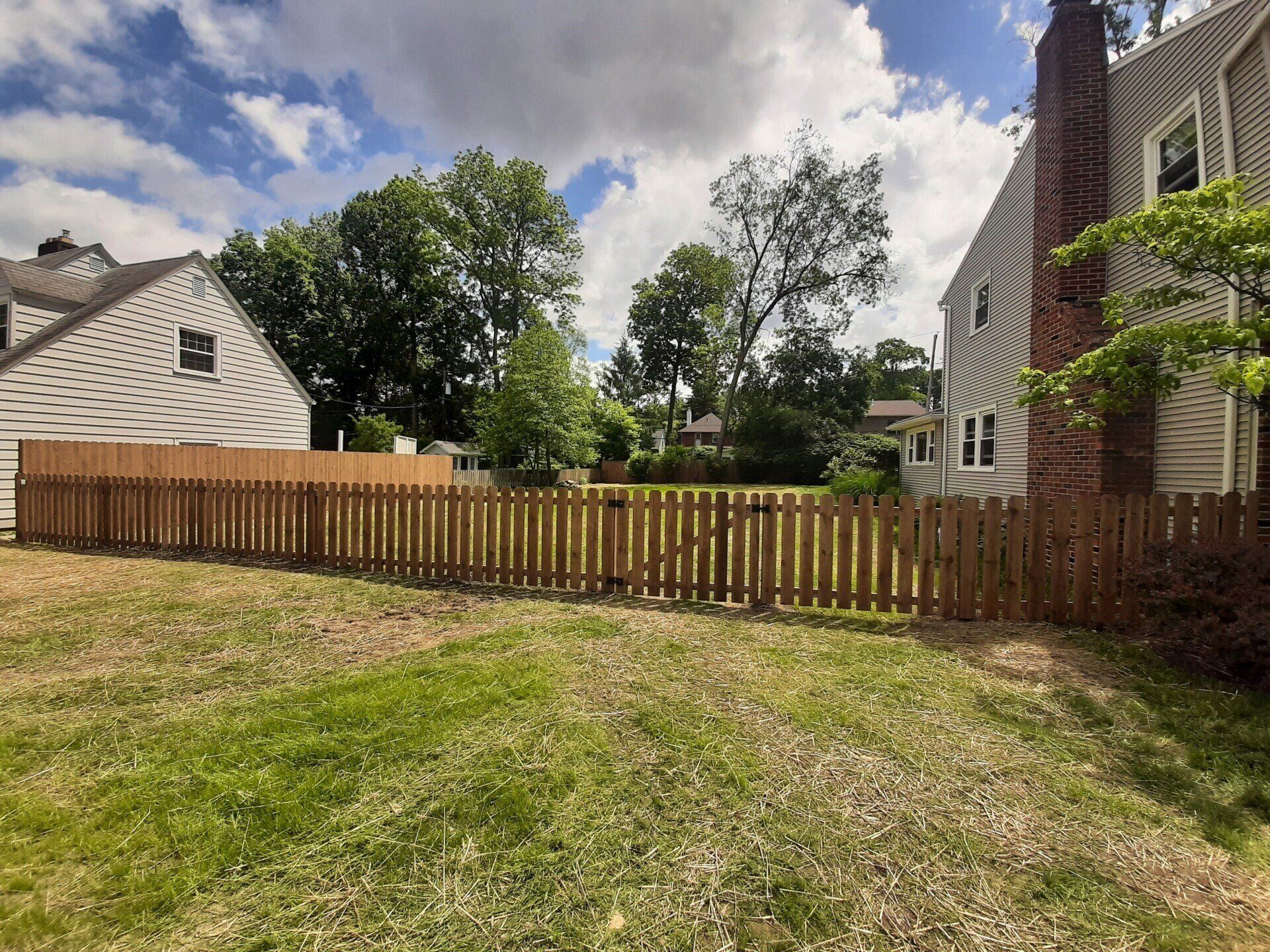 Brown wooden fence surrounding a grassy backyard with two houses in the background under a cloudy sky.