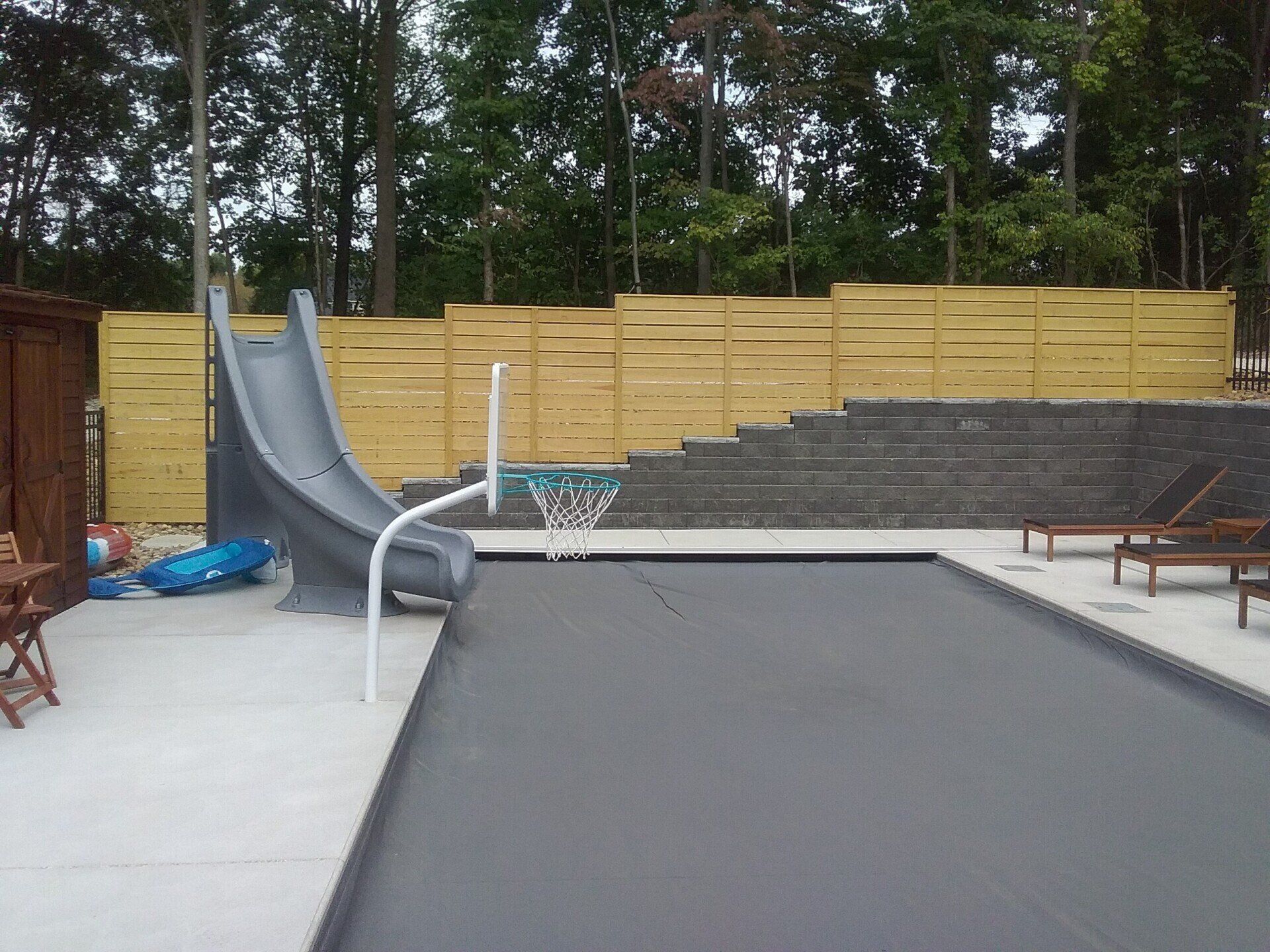 Outdoor pool with gray slide, basketball hoop, and dark pool cover. Wooden fence and trees in background.