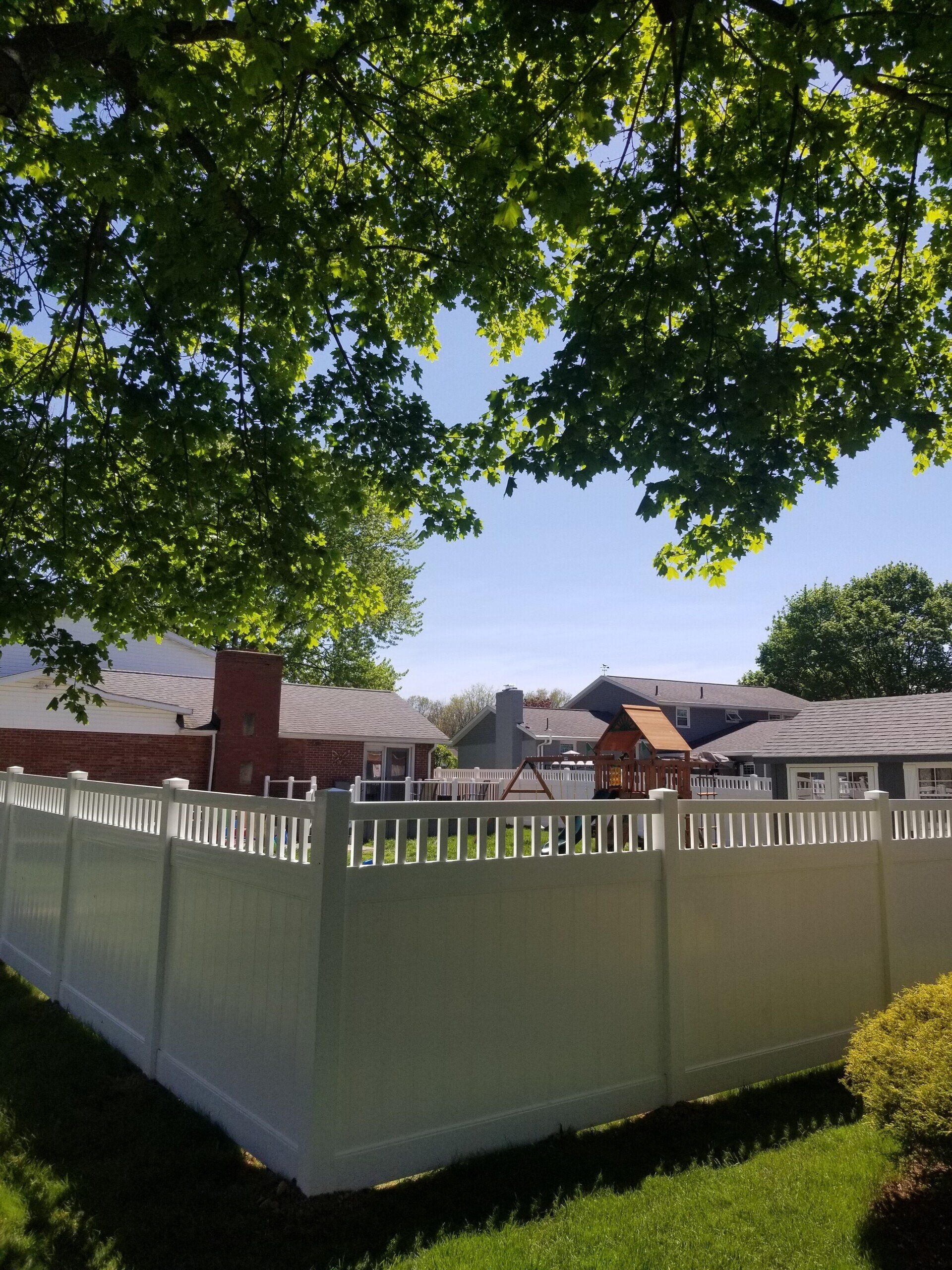 White fence in a yard, under tree branches. Houses visible in the distance under a blue sky.