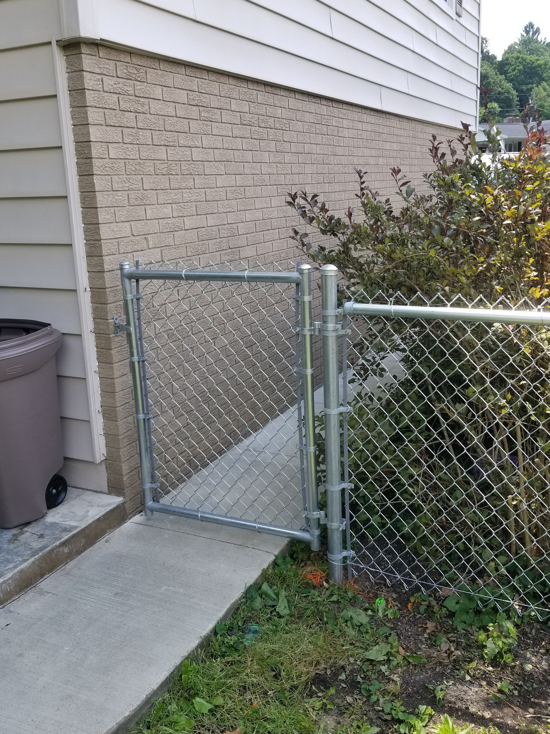 Chain link gate on a concrete path leading to a house, with a trash can on the left and bushes on the right.