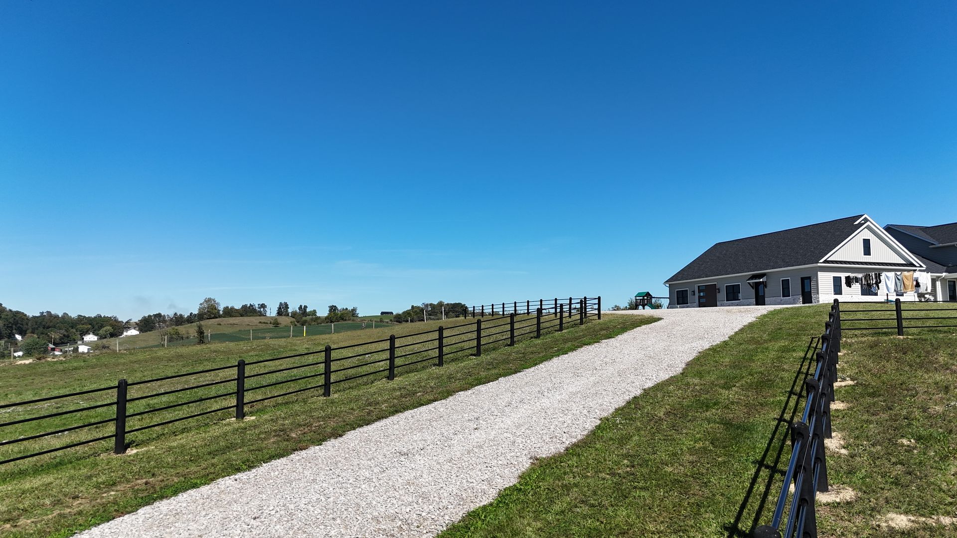 Gravel driveway leading to a white house with a dark roof on a grassy hill; black fence lines the drive; blue sky.