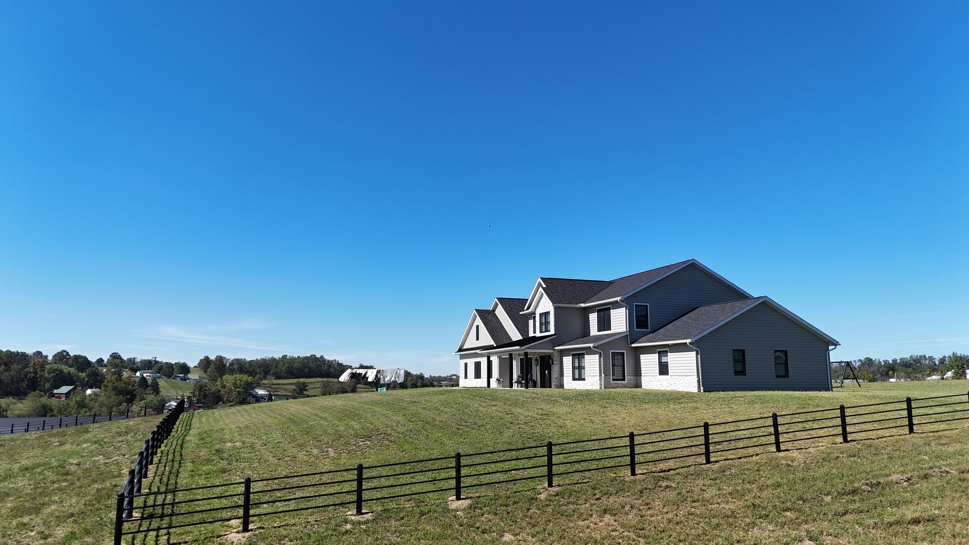 A two-story house on a grassy hill, black fence, and a clear blue sky.
