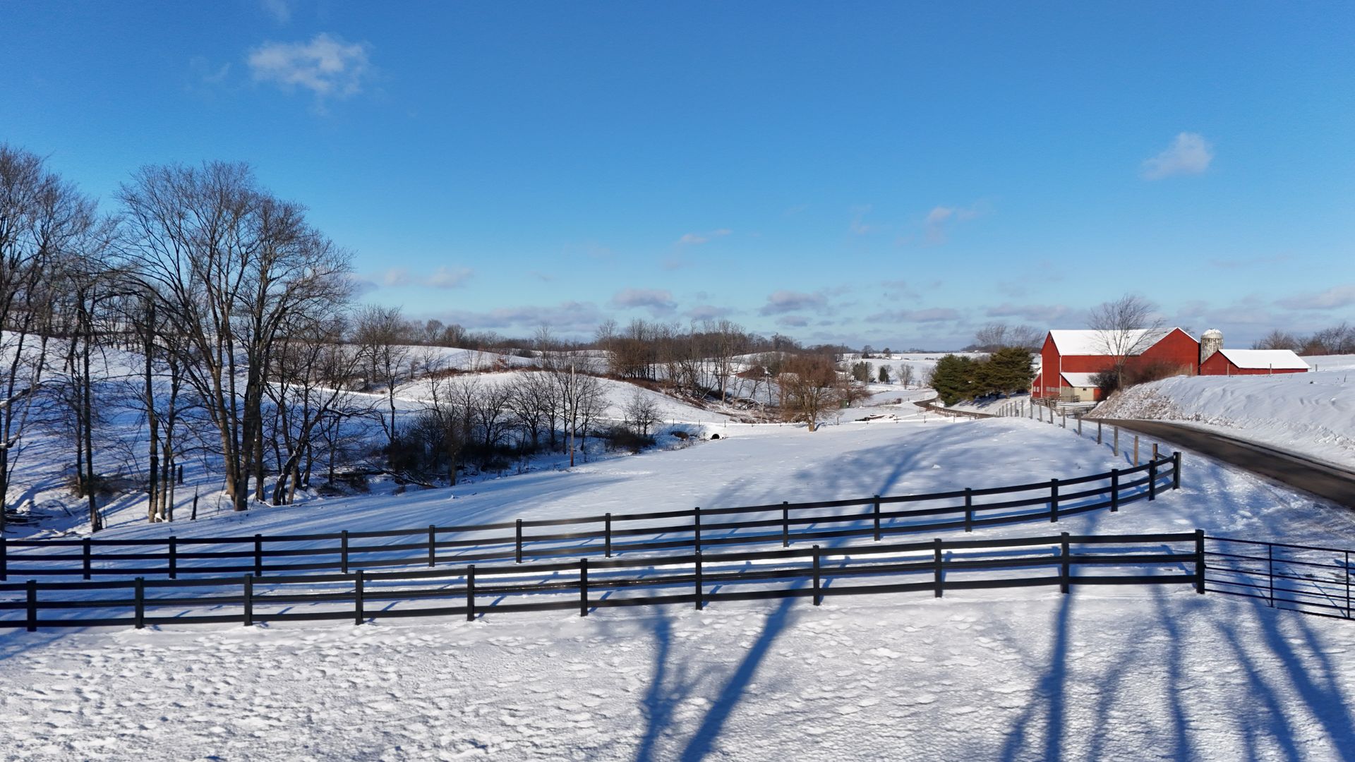 Snowy farm landscape with a red barn, trees, and black fence under a bright blue sky.