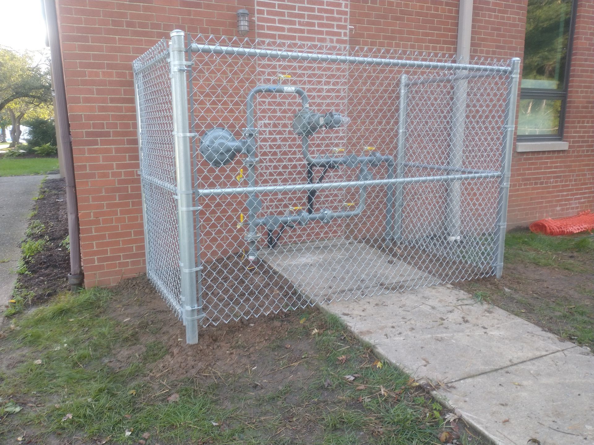 Chain-link fence enclosing gas meters near a brick building; concrete pathway in front.