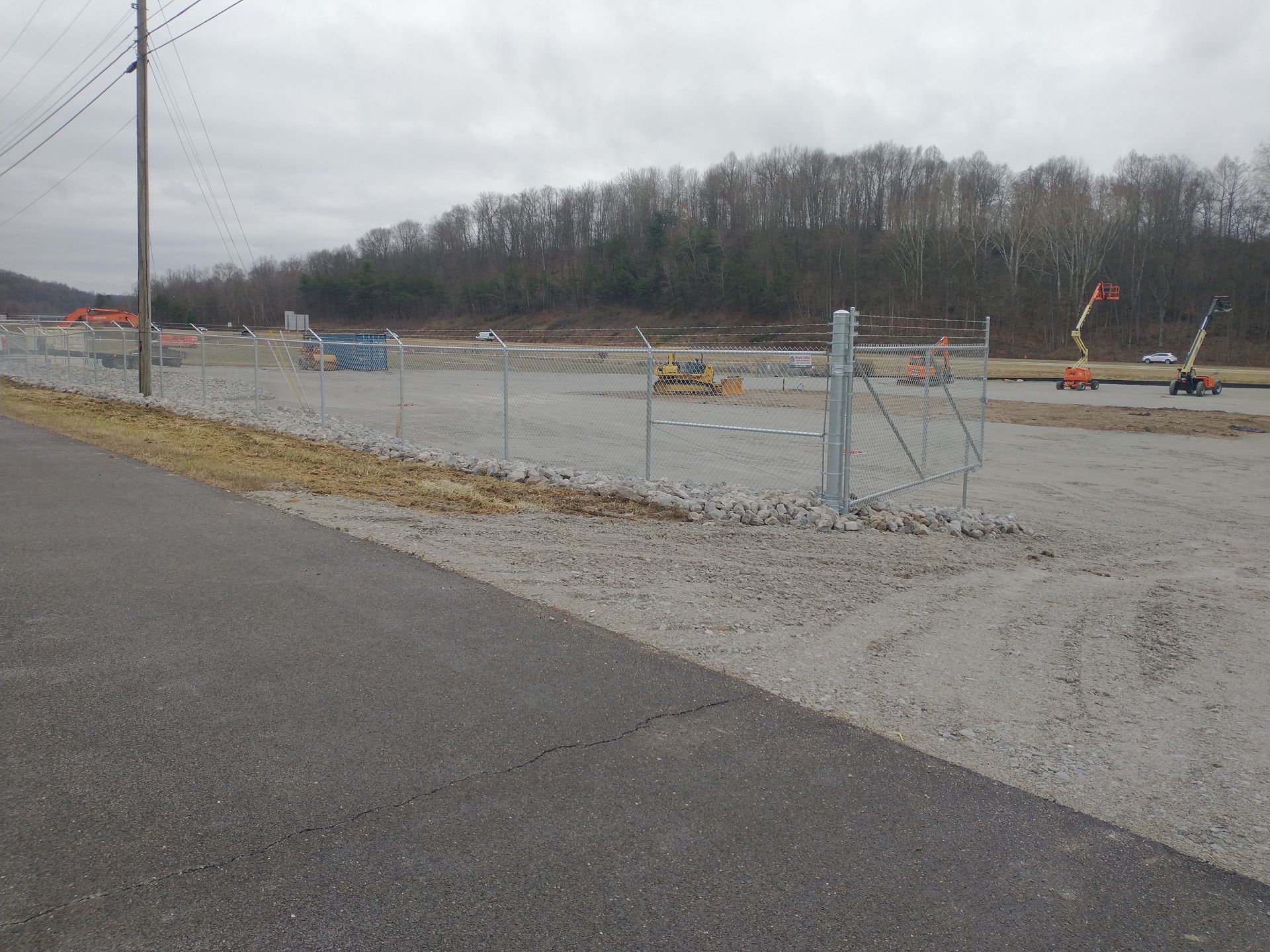 Chain-link fence surrounding a construction site; overcast sky, gravel ground. Trees in the background.