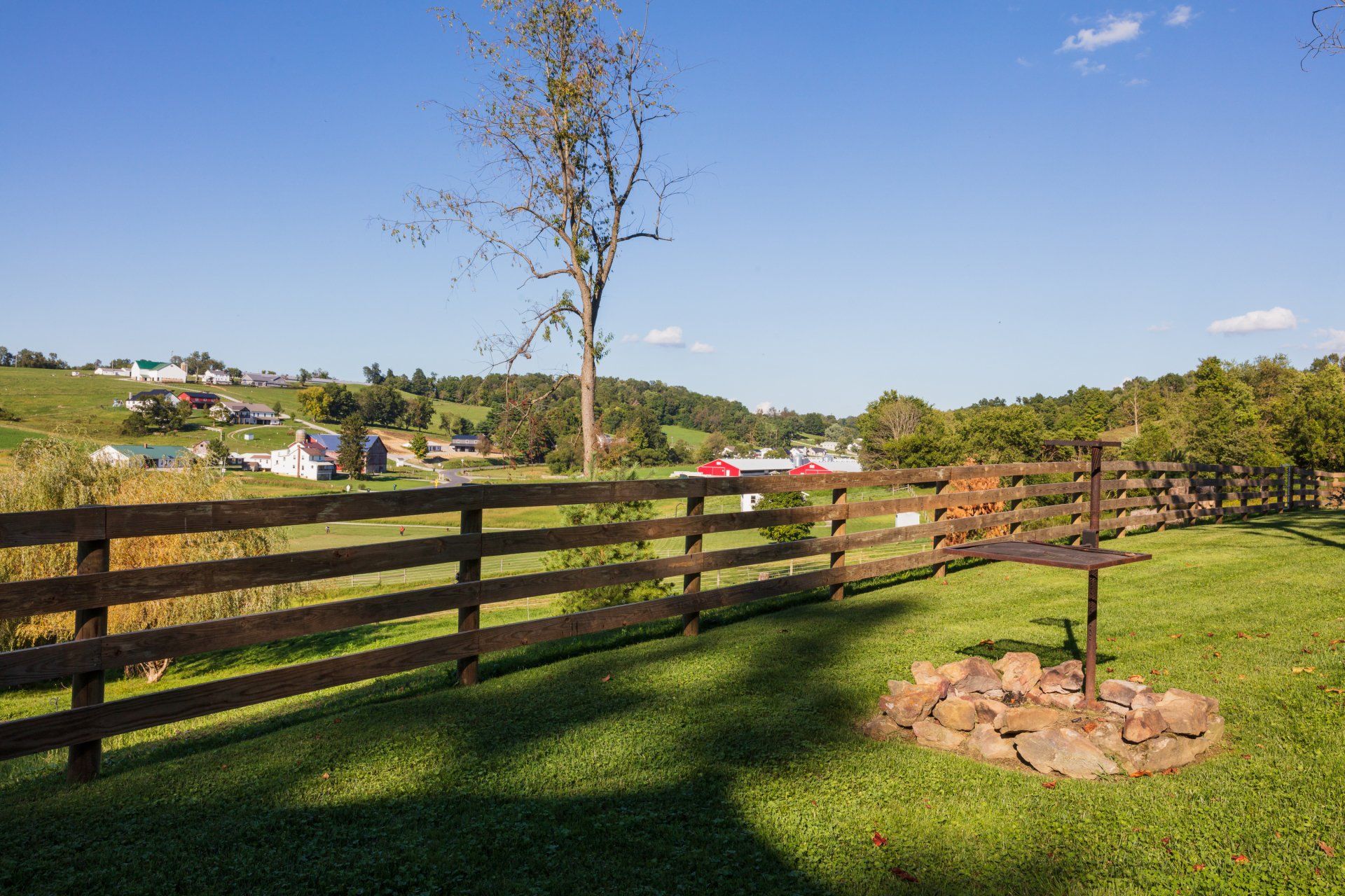 Wooden fence bordering a grassy field, with a fire pit and a distant view of houses and trees under a blue sky.