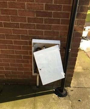 Open electrical panel on a brick wall with a white door hanging loose near a downspout.
